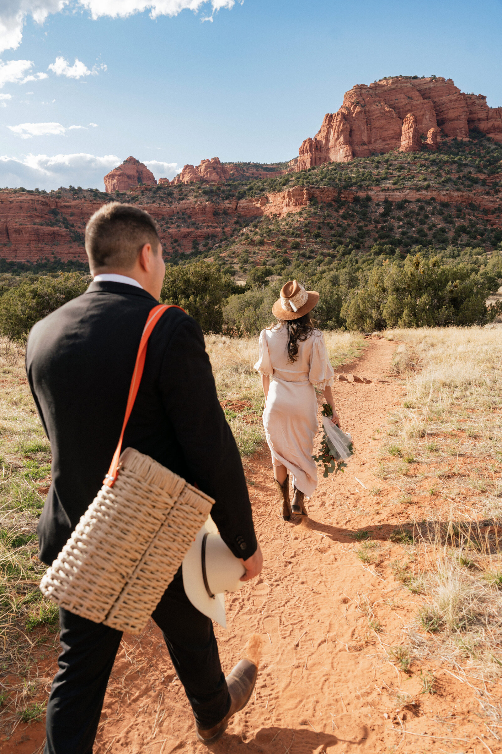 Bride and groom hike along a red dirt trail at Bear Mountain in Sedona, Arizona, during their western-inspired elopement with cowboy boots, a bouquet, and a picnic basket.