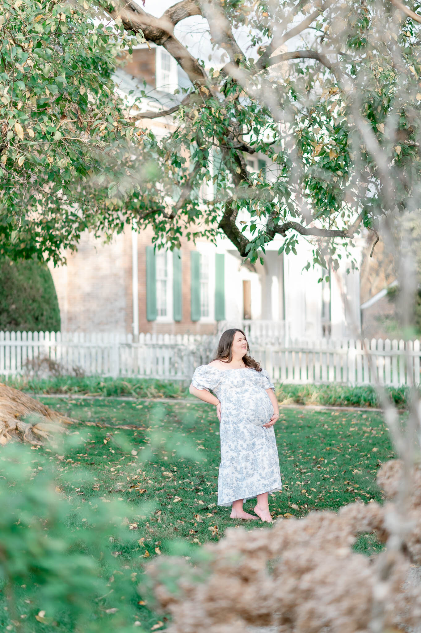 Pregnant woman stands below a large oak tree in a blue and white dress with fallen leaves on the ground