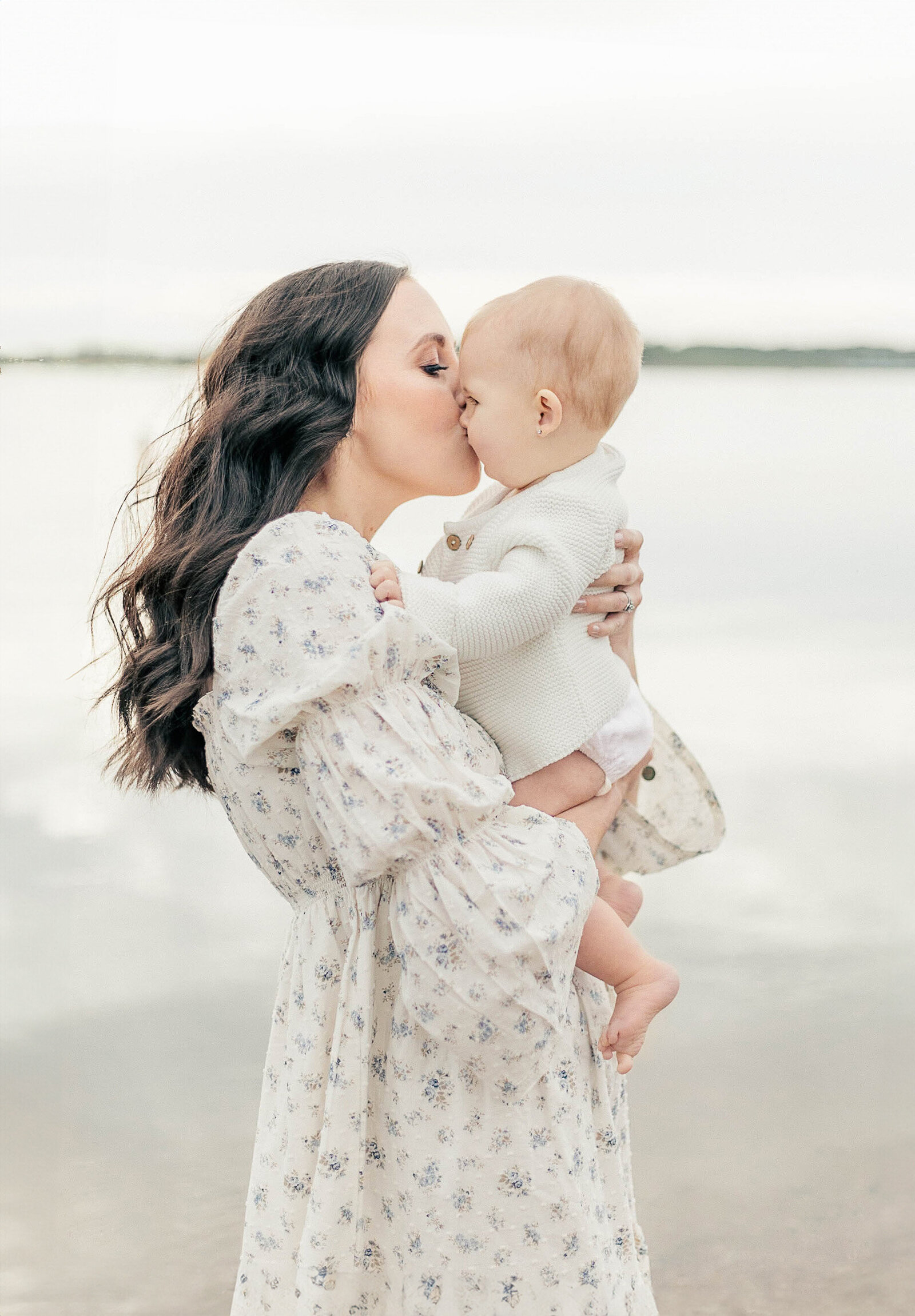 Mother in a floral dress kissing her baby while standing at the edge of a calm lake in Kansas City.