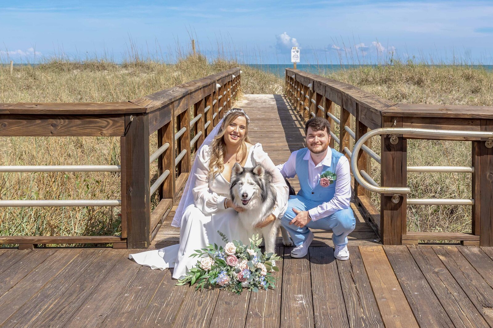 The bride and groom sit with their fluffy dog on a wooden boardwalk leading to the beach, captured at a Carolina Beach, NC wedding, highlighting a pet-inclusive and coastal wedding day.