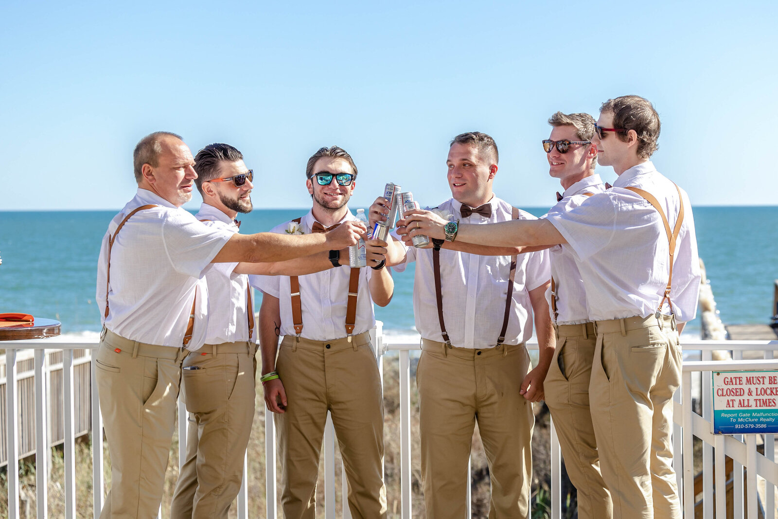 A group of groomsmen dressed in khakis and suspenders toast with drinks on a beachside balcony during a wedding in Wrightsville Beach, NC, blending coastal charm with camaraderie.