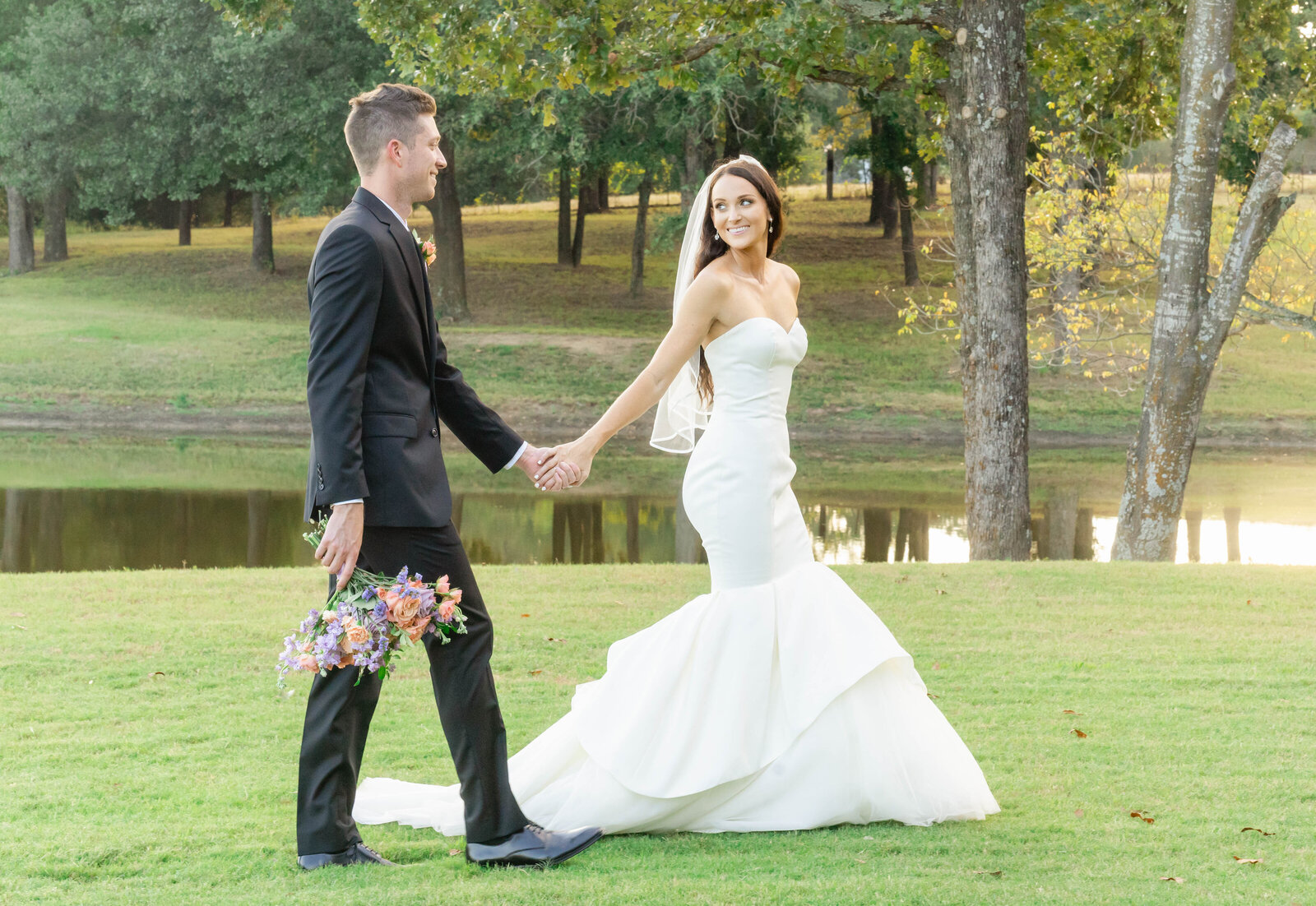 Portrait of a bride and groom in a white wedding gown and black tuxedo with a bouquet of flowers walking on grass.