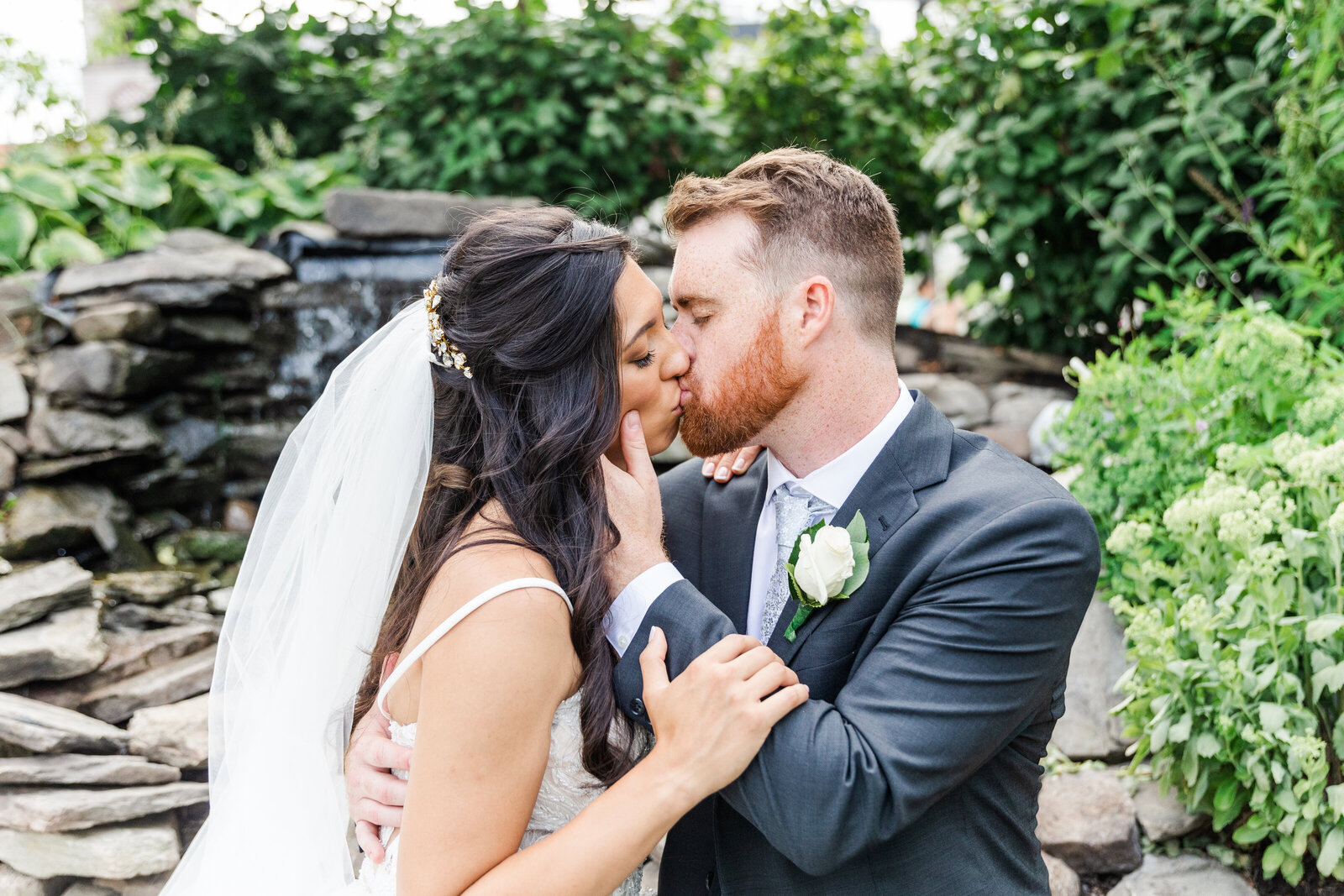 bride and groom sit on the rocks of a waterfall in ohio kissing on their wedding day. photographed by toledo based wedding photographer.