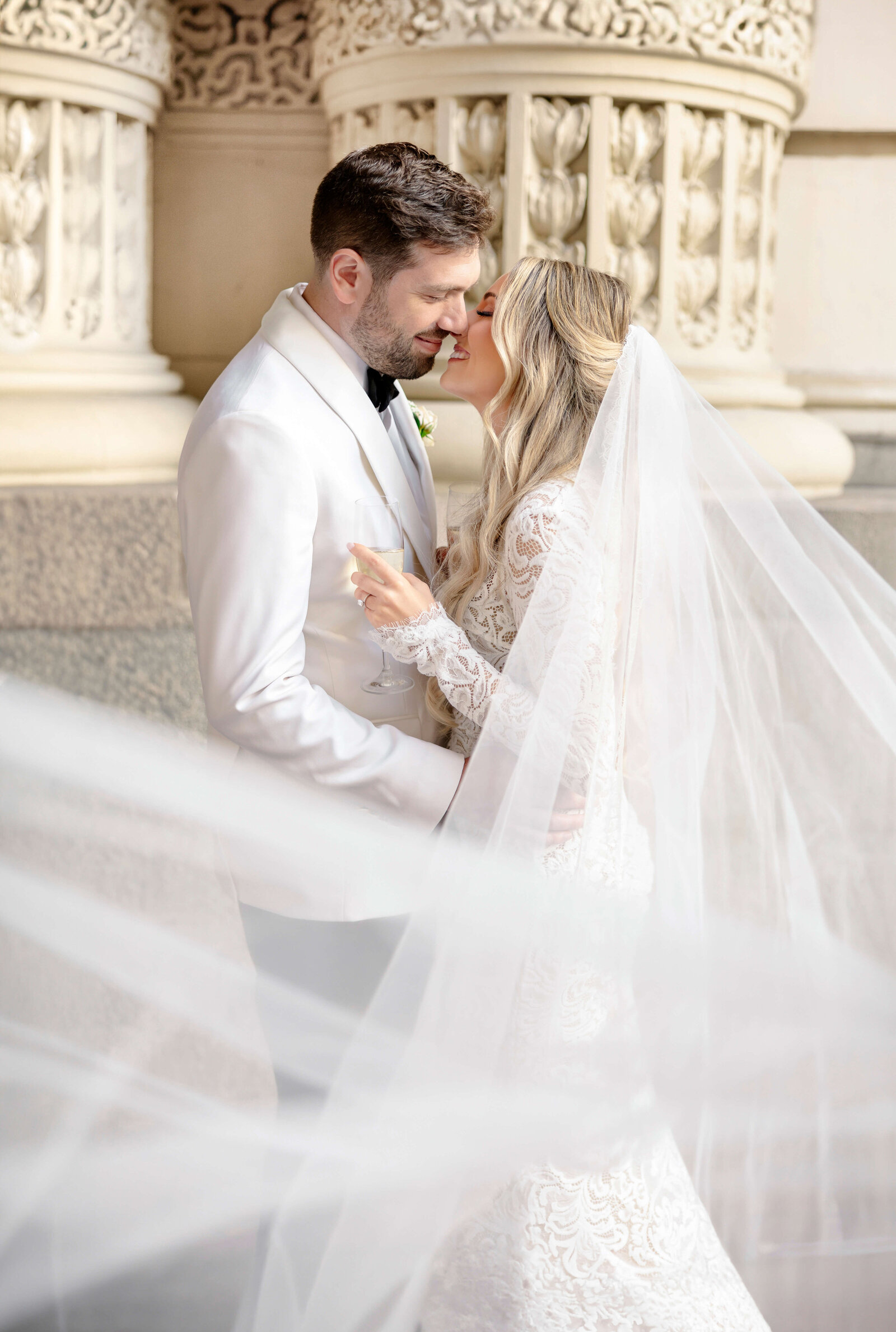 A bride and groom in elegant attire share an intimate moment. The bride wears a long lace veil that flows in the foreground, while the groom is in a white tuxedo jacket. Theyre standing close, smiling at each other near a carved stone wall.