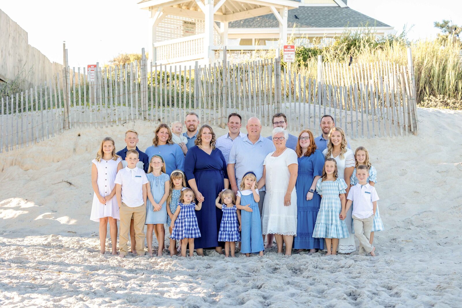 A large family stands together on sandy dunes with a beach house in the background, dressed in coordinating blue and white outfits. The scene captures a classic coastal family moment, ideal for destination family photographer NC, showcasing unity and elegance.