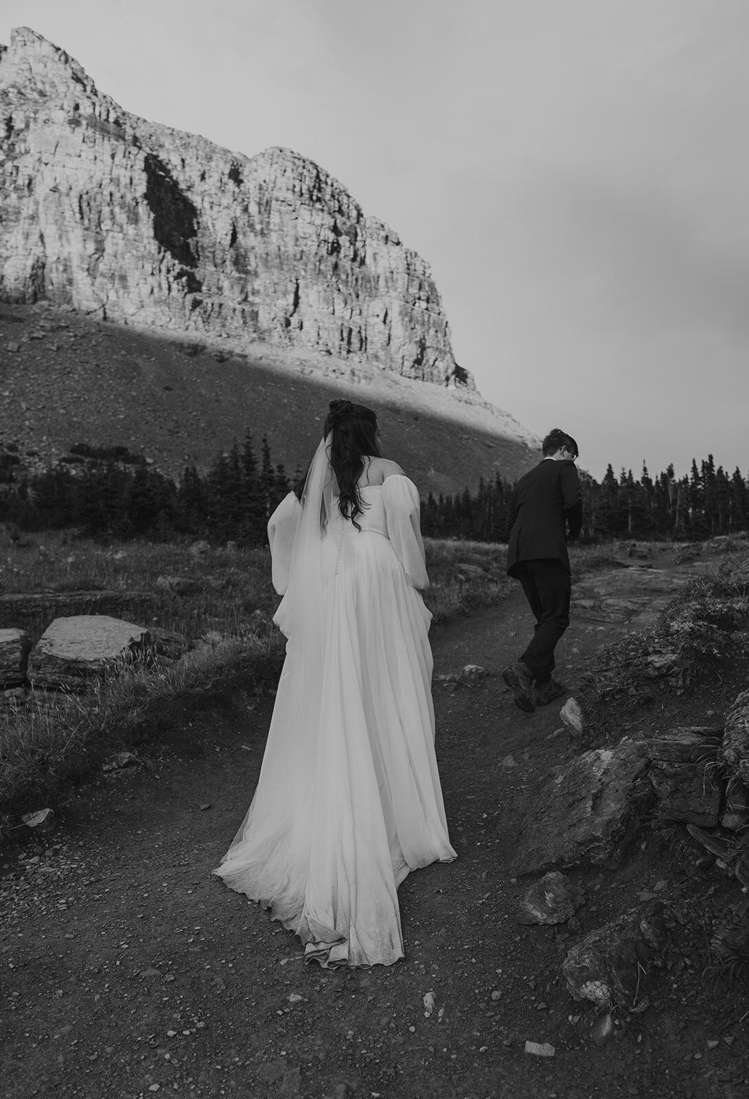 A bride follows her groom up a rugged mountain trail surrounded by cliffs and evergreens during their Glacier National Park elopement, captured in black and white by Sydney Breann Photography.