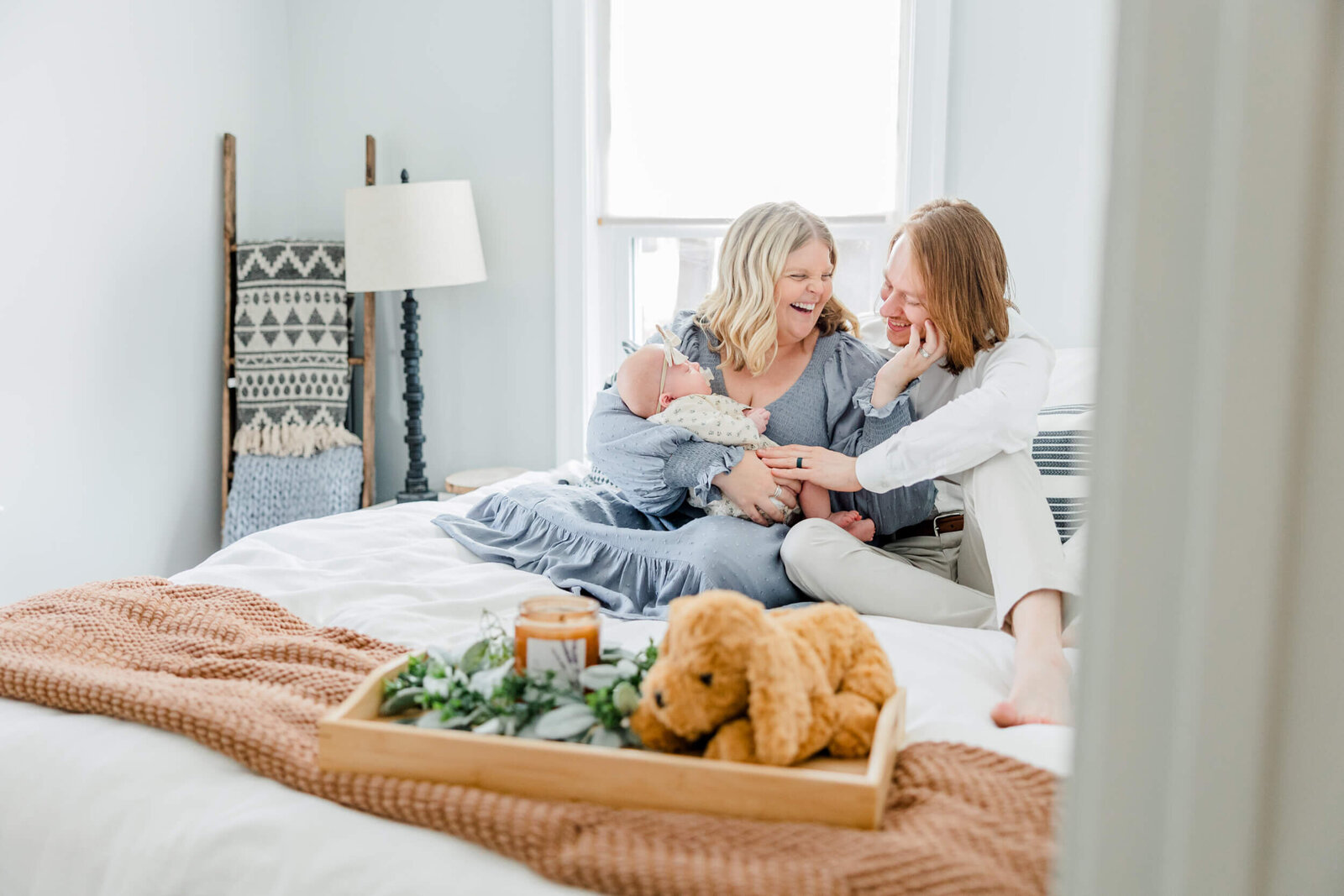 Mom and dad laugh together on the bed while mom holds a newborn