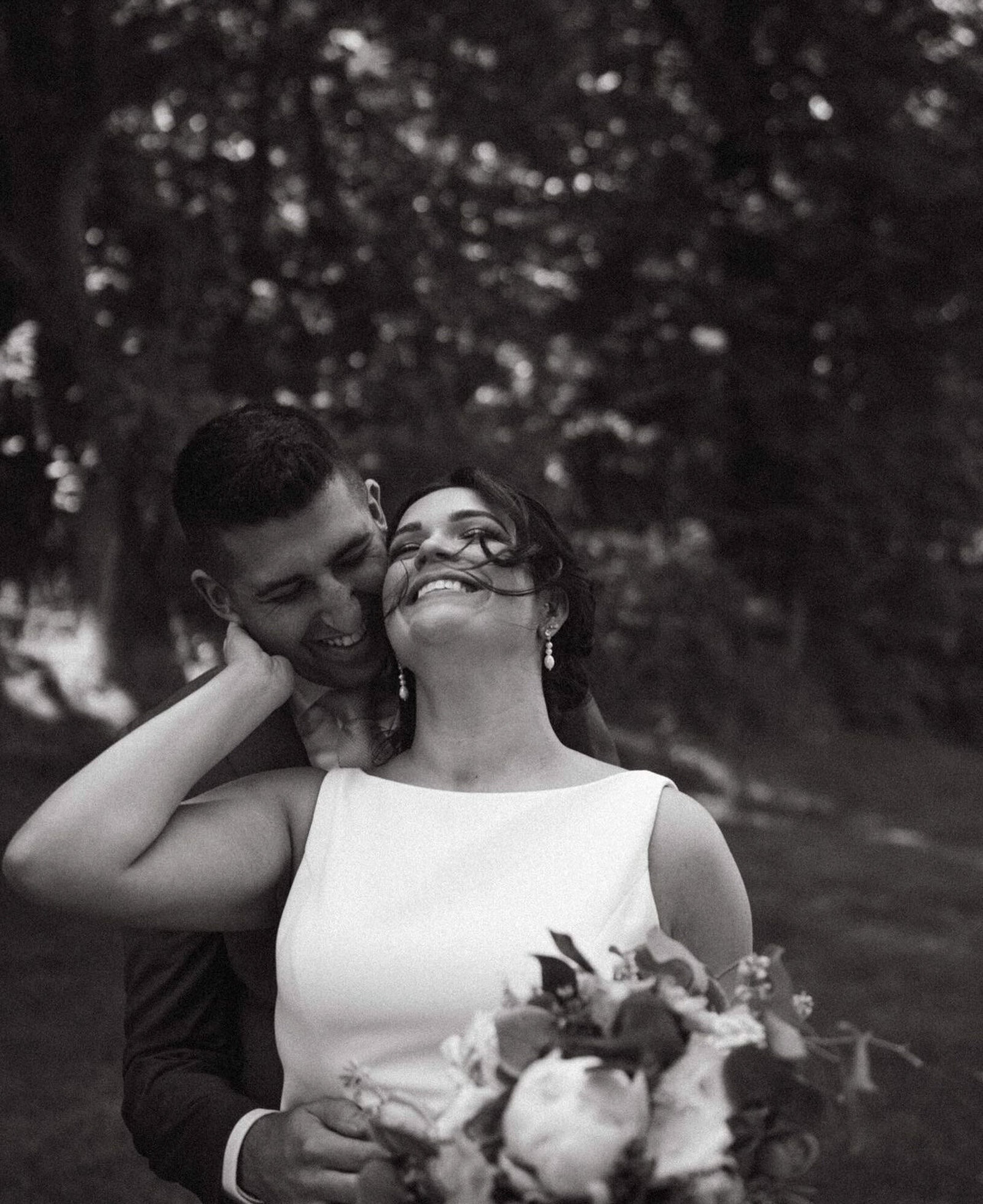 black and white photo of groom embraces the bride from behind, both smiling deeply and bride holds a bouquet, both standing in a lush, tree-filled setting