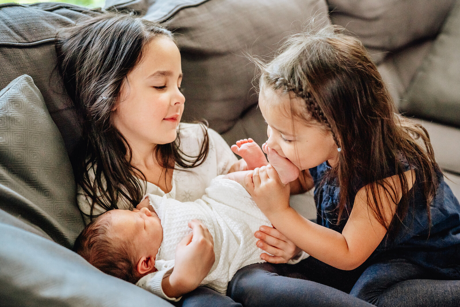 Two young sisters sit on a couch, lovingly gazing at their newborn sibling. One sister holds the baby while the other playfully kisses the baby’s tiny feet.