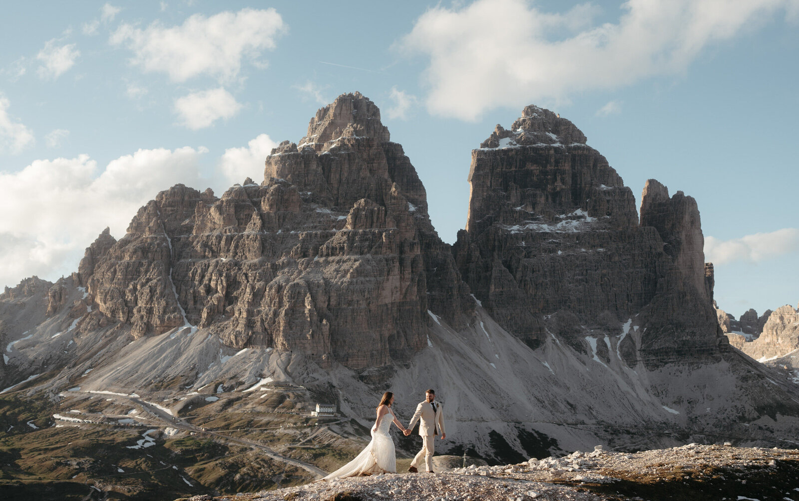 Couple hikes hand in hand during their hiking wedding in the Dolomites, there is a beautiful mountain view behind them.