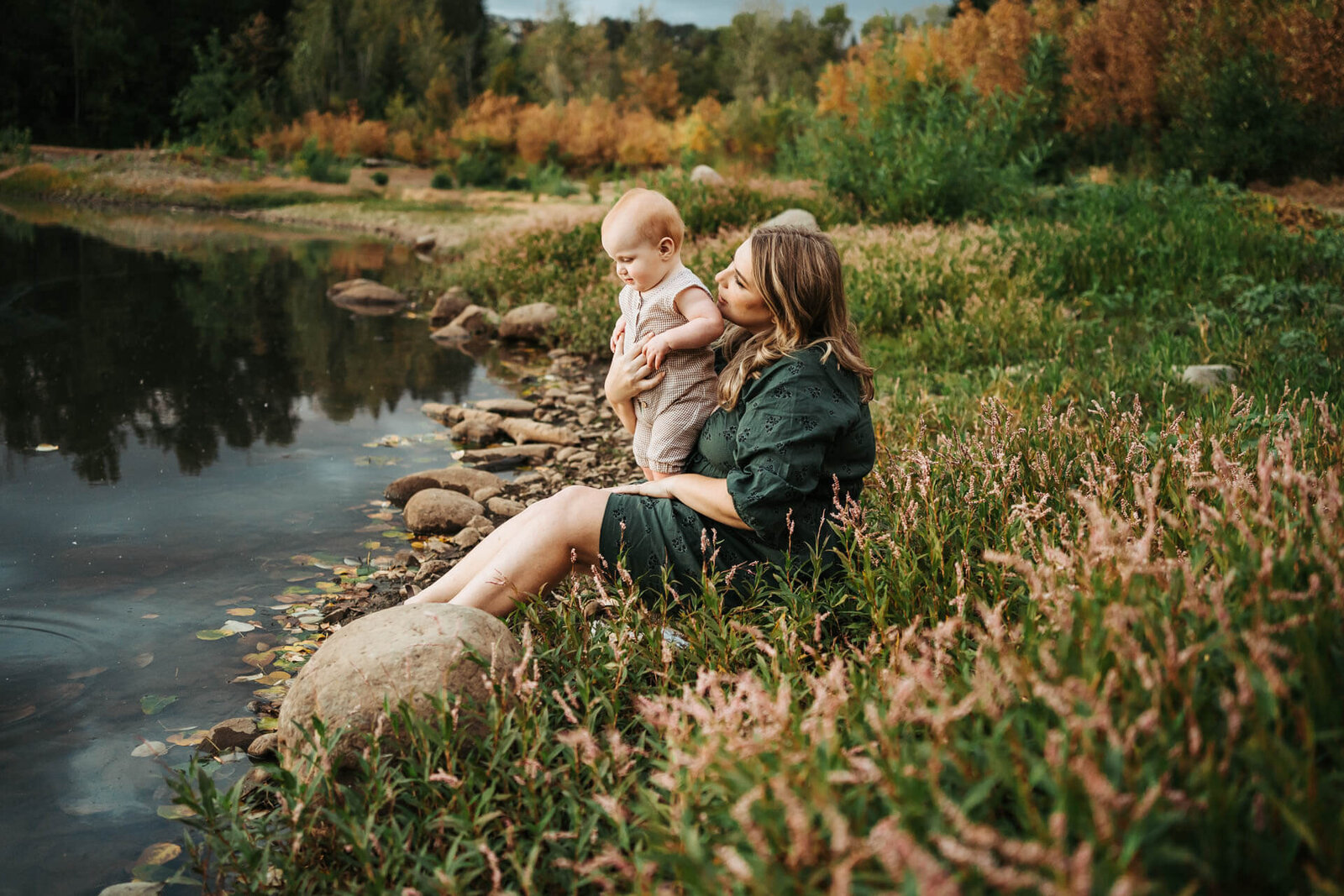Mom in green dress holding her baby on her lap and looking at the pond