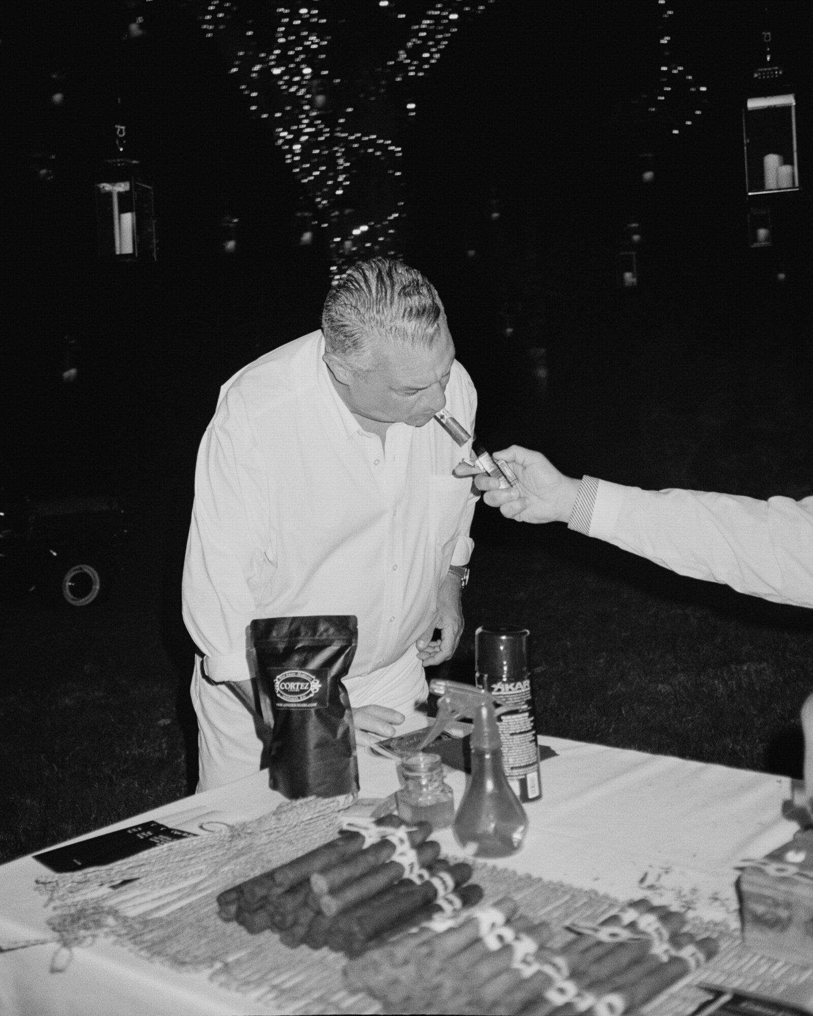 Candid black & white documentary style direct flash photo of cigar rollers arm reaching into frame to light a man's cigar 