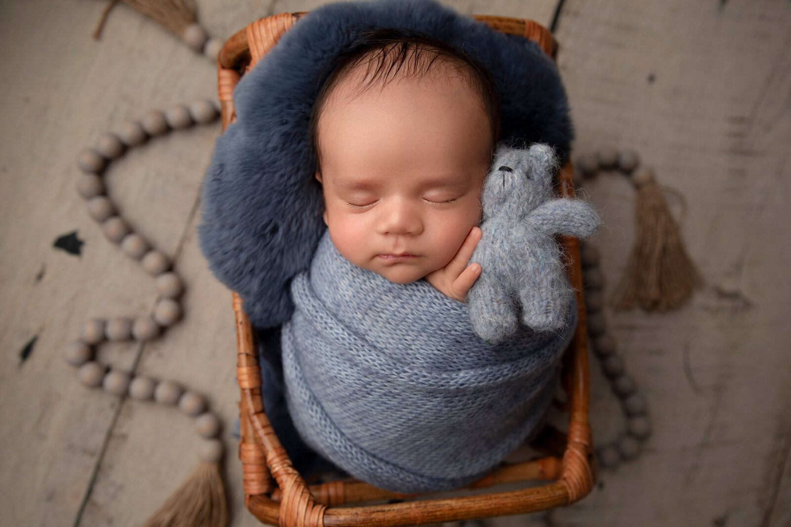 A peaceful newborn baby sleeps snugly in a basket adorned with soft blue pastel blankets, gently holding a tiny teddy bear. Captured at Little Legacies Studio in Phoenix, Arizona, this heartwarming scene showcases the innocence and charm of your baby's earliest days.