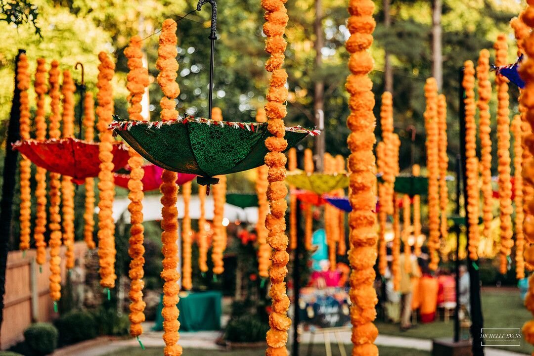 Vibrant Indian wedding decor featuring marigold garlands and colorful hanging umbrellas at an outdoor celebration, designed for a traditional mehndi or sangeet event with festive cultural elements.