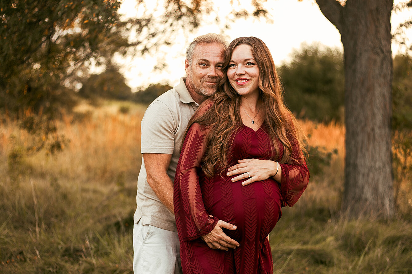 Meadow at sunset. Man and woman in the center of picture facing the camera smiling. He is standing behind her. Heads are leaning into each other both are embracing her maternity belly. Maternity Photographer.