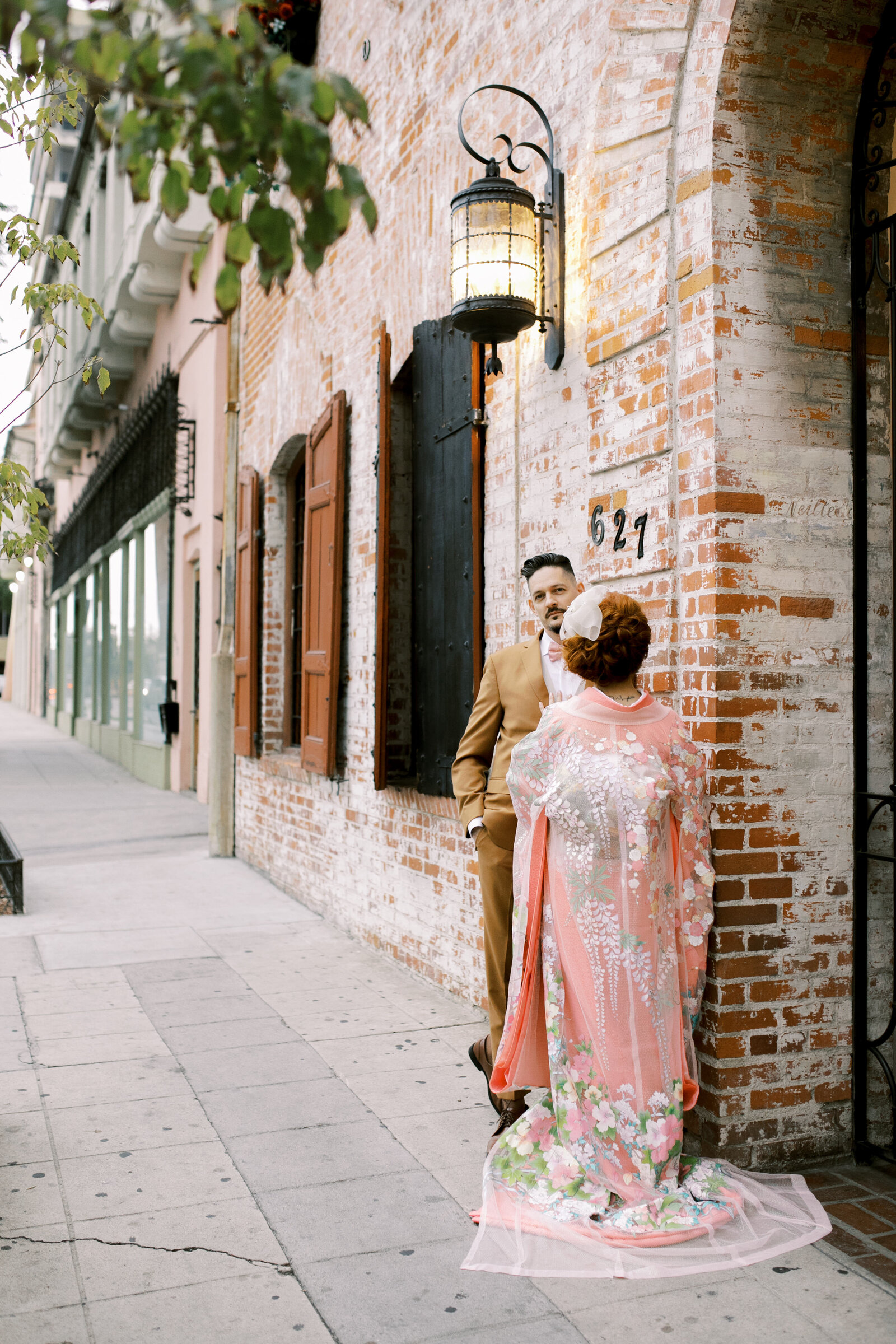 Kimono rental Los Angeles – Bride in pink iro-uchikake faces groom beneath vintage lantern along the brick exterior of Carondelet House | ロサンゼルス カロンデレットハウス外観、ヴィンテージランタン下で着物レンタル花嫁と新郎が見つめ合う