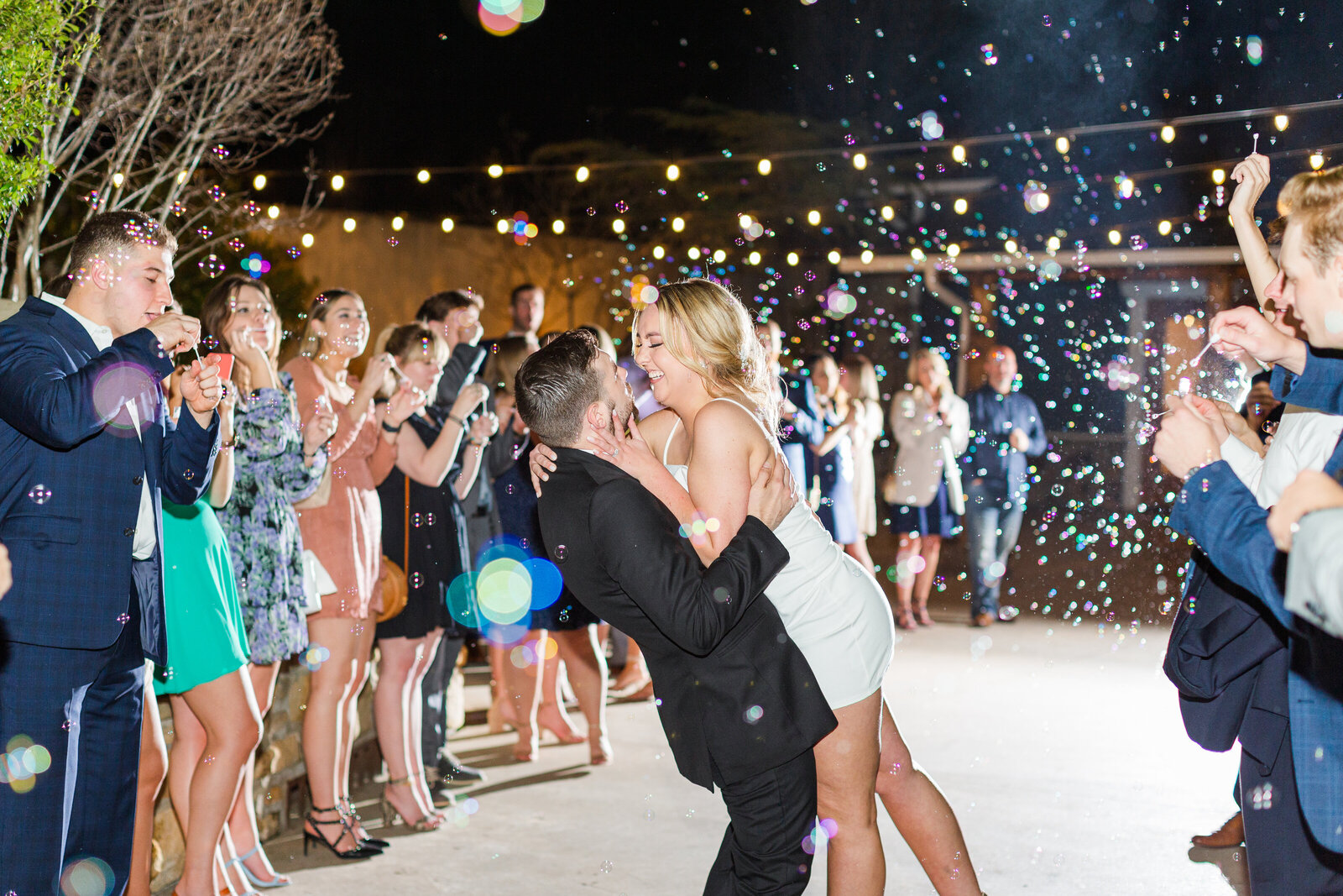 Couple kisses as they exit their luxurious wedding at Silo Event Center while guests cheer and blow bubbles.