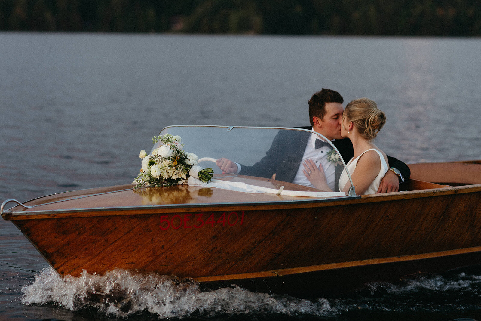 A couple kisses while seated in a small wooden boat adorned with flowers, drifting serenely across the water—a scene beautifully captured by Muskoka wedding photographer, Jennifer van Son.