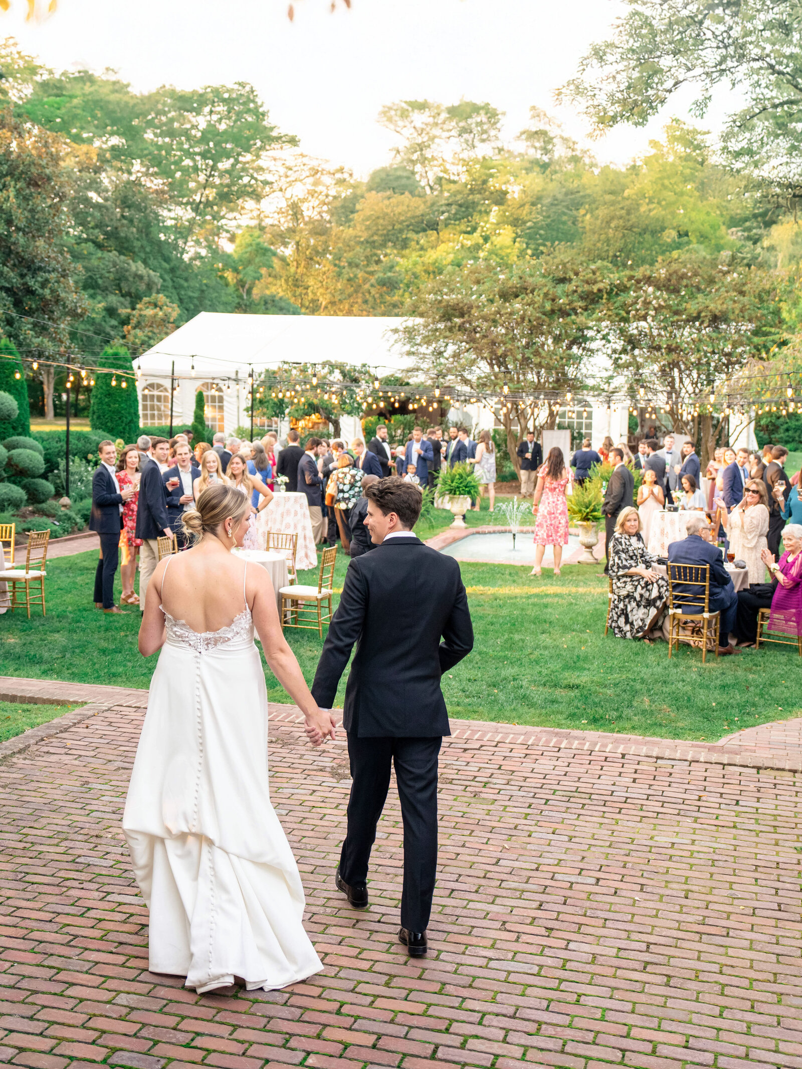 A bride in a white gown and a groom in a black suit walk hand-in-hand toward a group of formally dressed guests at an outdoor wedding reception, with greenery and a tent in the background under a clear sky.