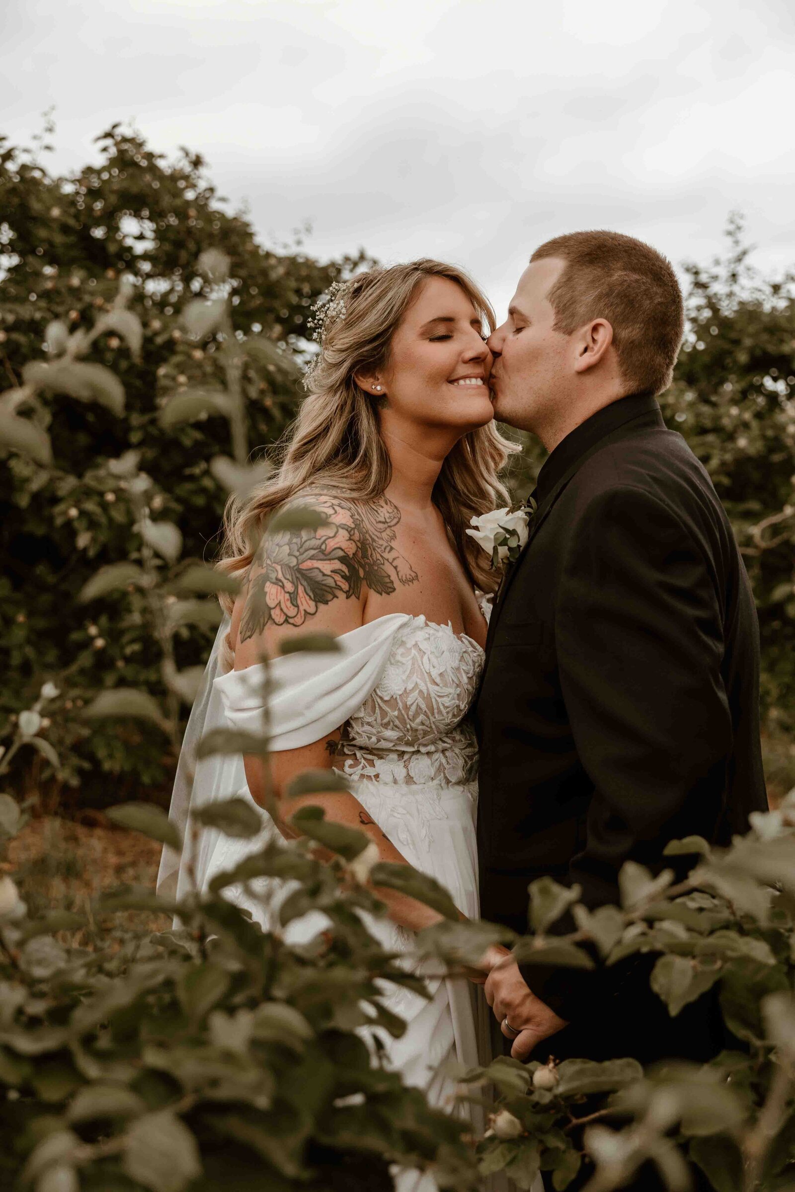 Married couple kissing on cheek between trees.