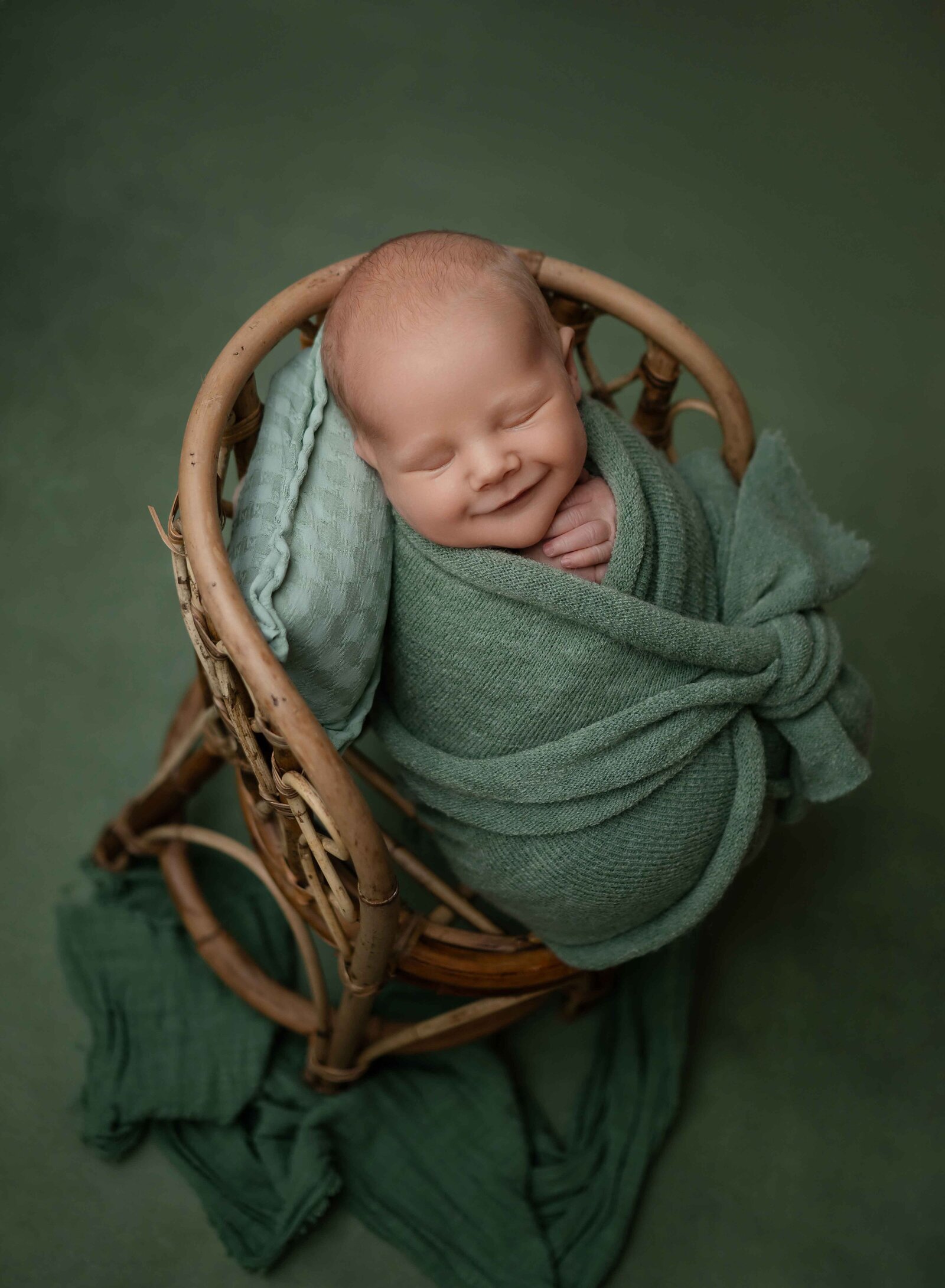 Baby in a green swaddle on a bamboo chair and smiling in his sleep.