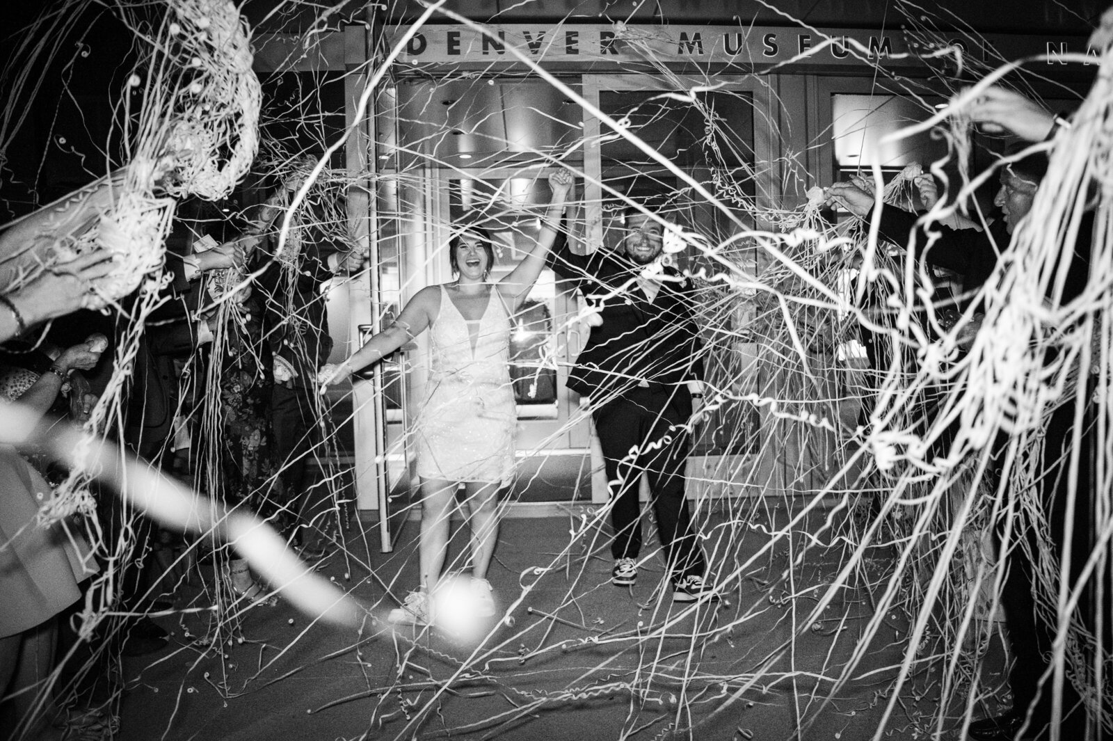 A bride and groom exit their wedding reception as their guests throw streamers in celebration.