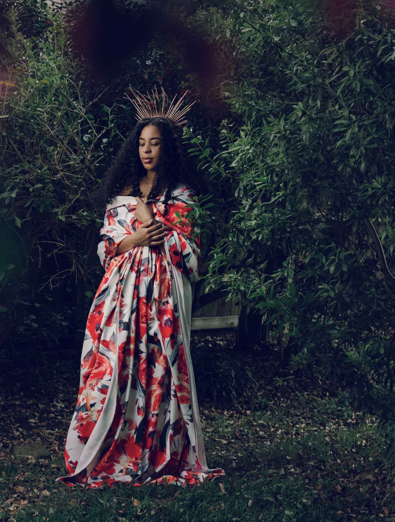 Darkly lit portrait of a woman in a floral dress, standing in front of trees, looking camera left. Houston's premier portraits.