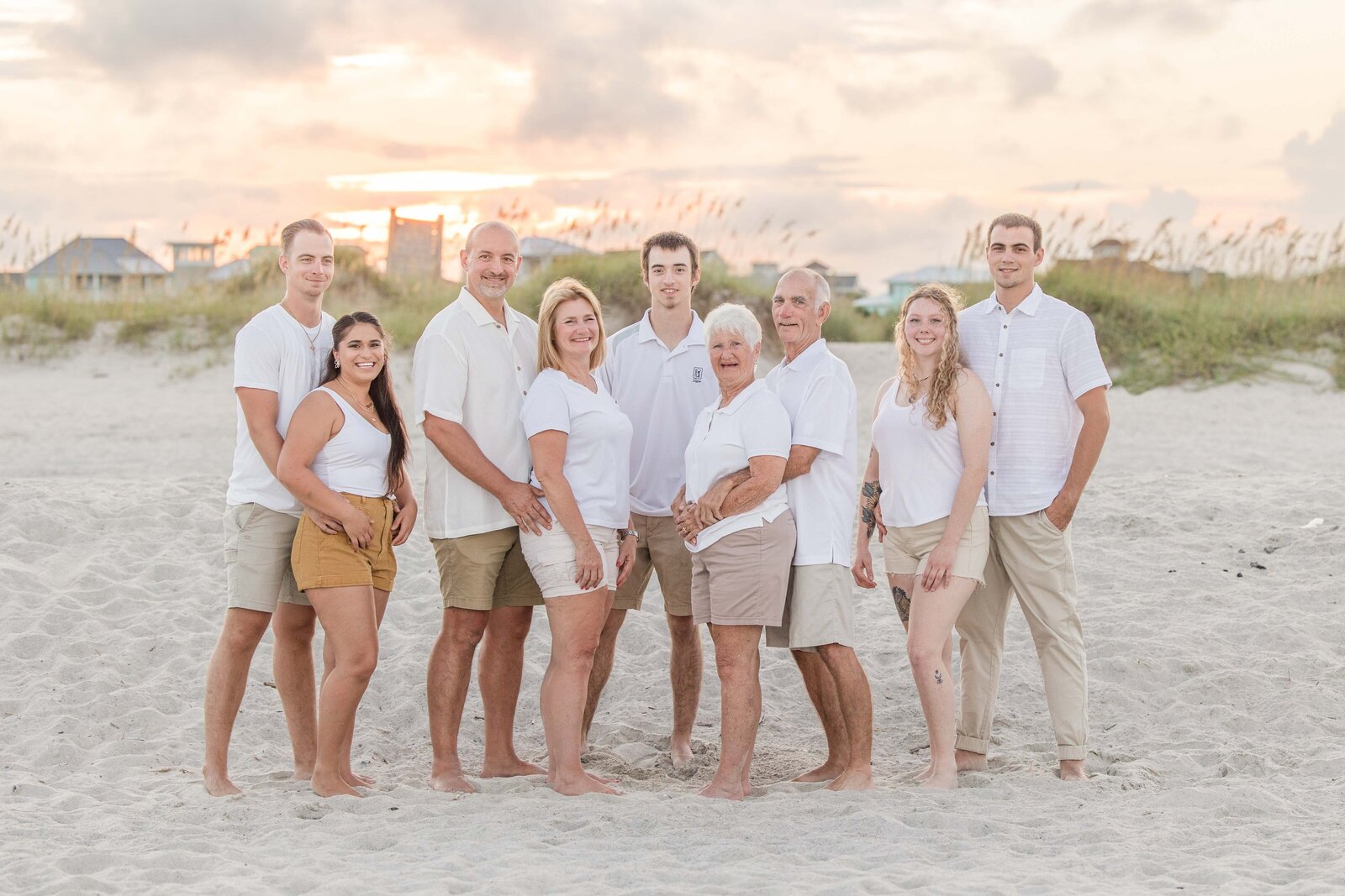 An extended family poses barefoot on the sandy shores of Ocean Isle Beach during a stunning sunset. Dressed in coordinated white and beige attire, they stand together, capturing the warmth of a family vacation. This image highlights professional family photography in NC, perfect for memorable family gatherings.