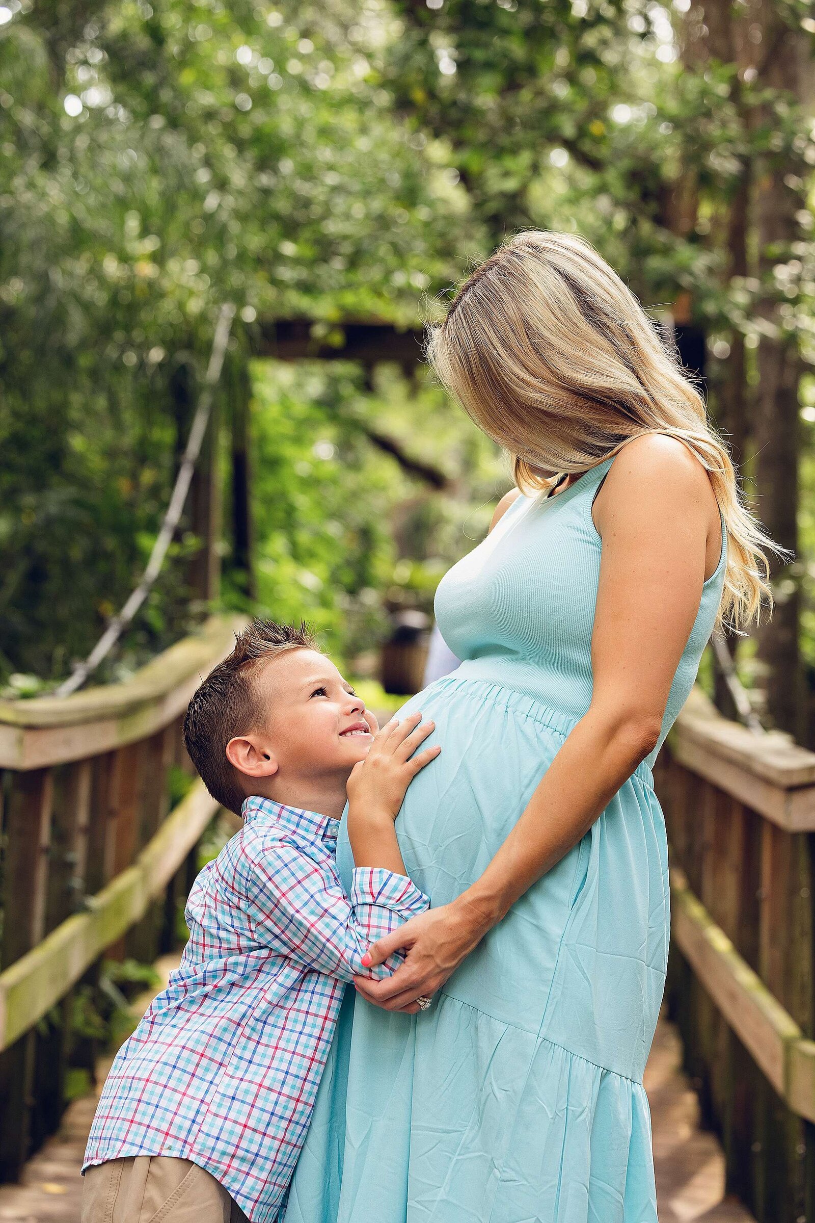 A joyful pregnant woman and her son pose on a wooden bridge, beautifully photographed by Bia Schaefer in Orlando.