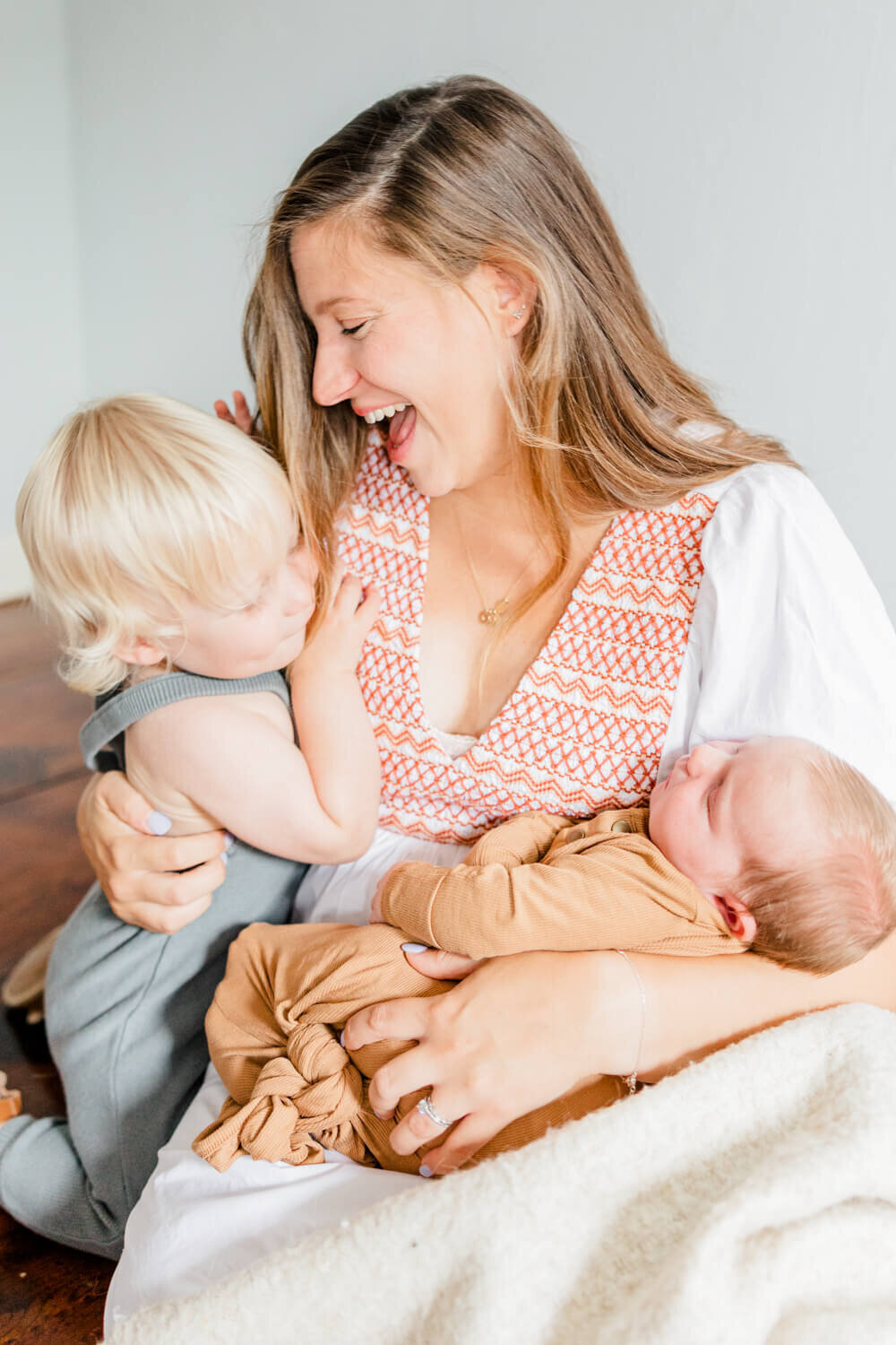 Mom sits on the floor, holding newborn in one arm and hugging her toddler in the other