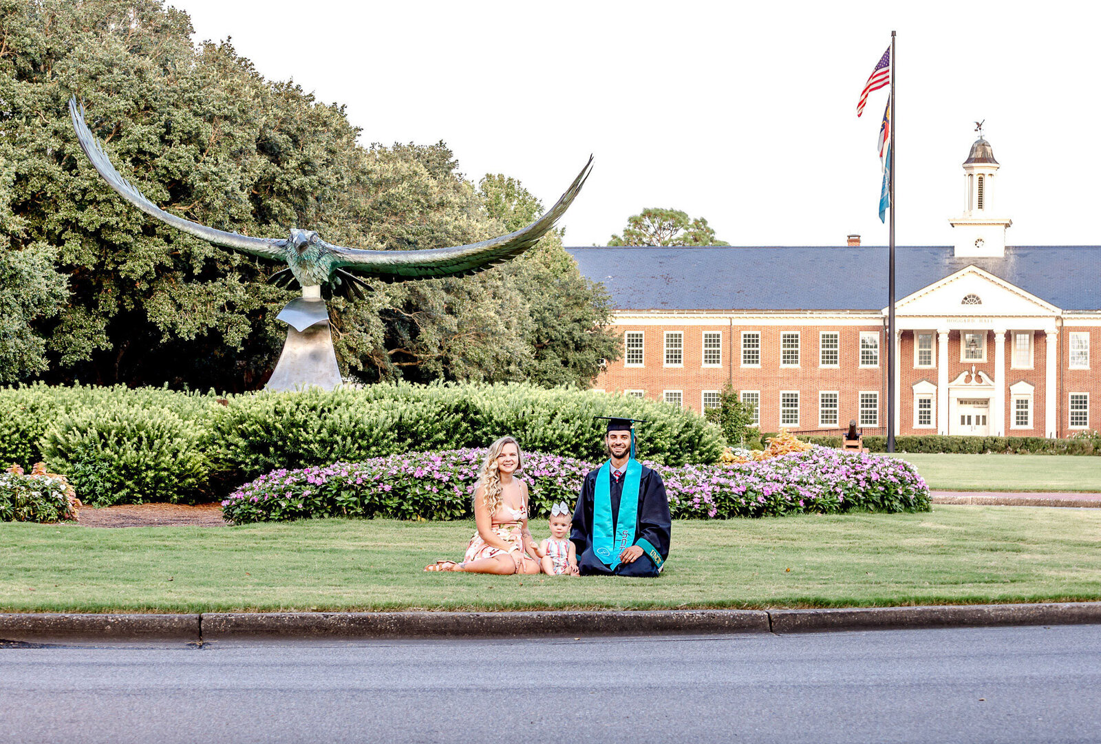 A college graduate in academic regalia sitting on the lawn with his family, with the seahawk statue and a University of North Carolina Wilmington building in the background in Wilmington, North Carolina. This photo captures the joy of graduation, perfect for those looking for memorable senior photography.