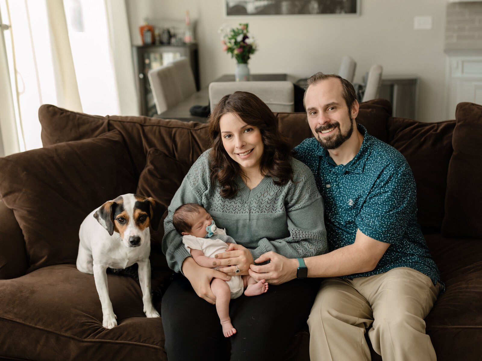 parents holding newborn baby boy sitting on couch with dog