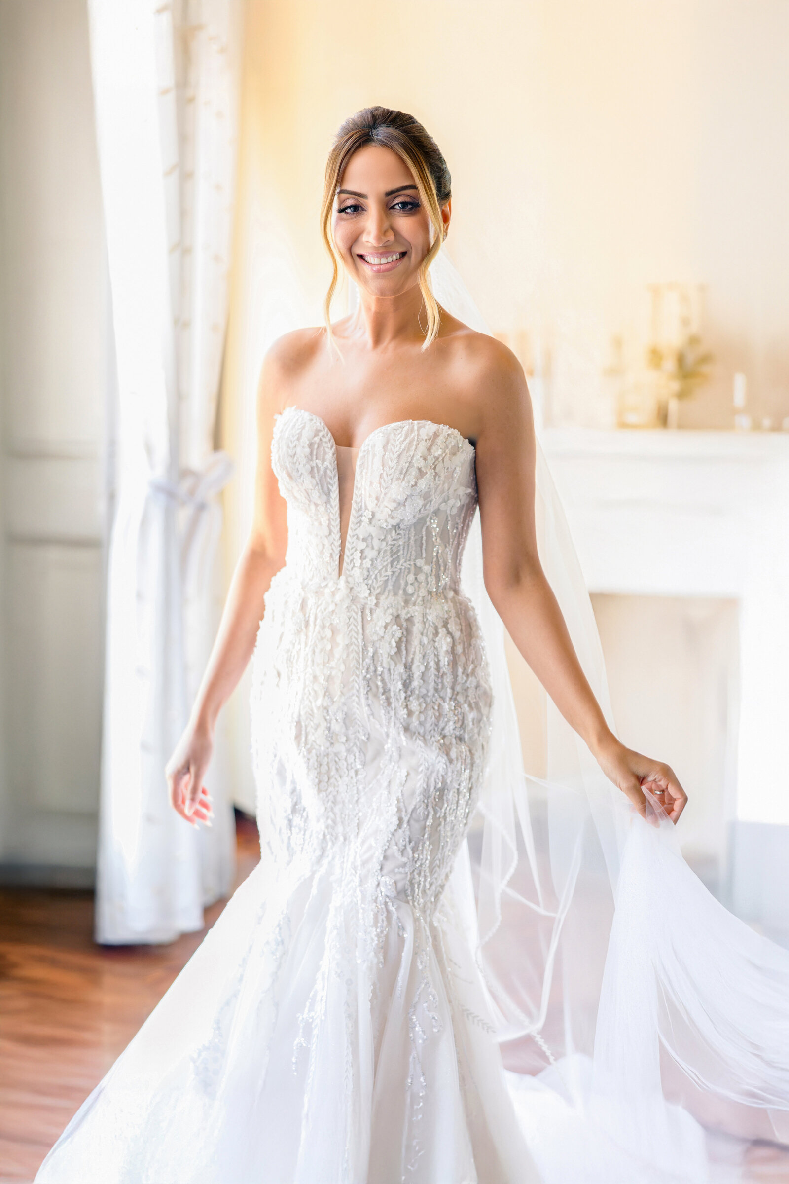 A woman in a strapless white wedding dress stands smiling indoors. The dress features intricate lace details and a long train. She holds part of the train while posing in a softly lit room with wooden floors and light-colored curtains.