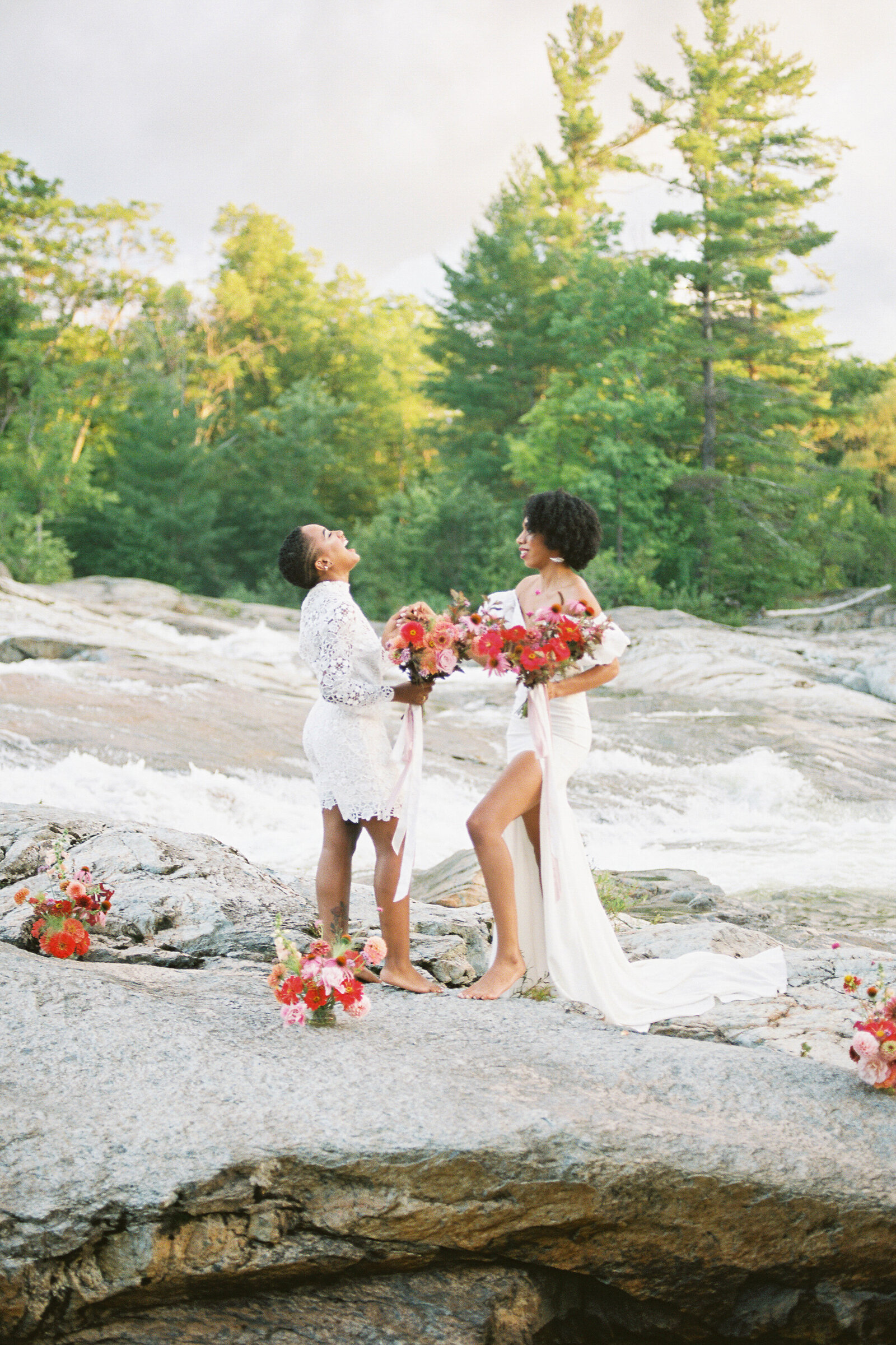 Two brides laughing while standing on rocks with flowers around them.
