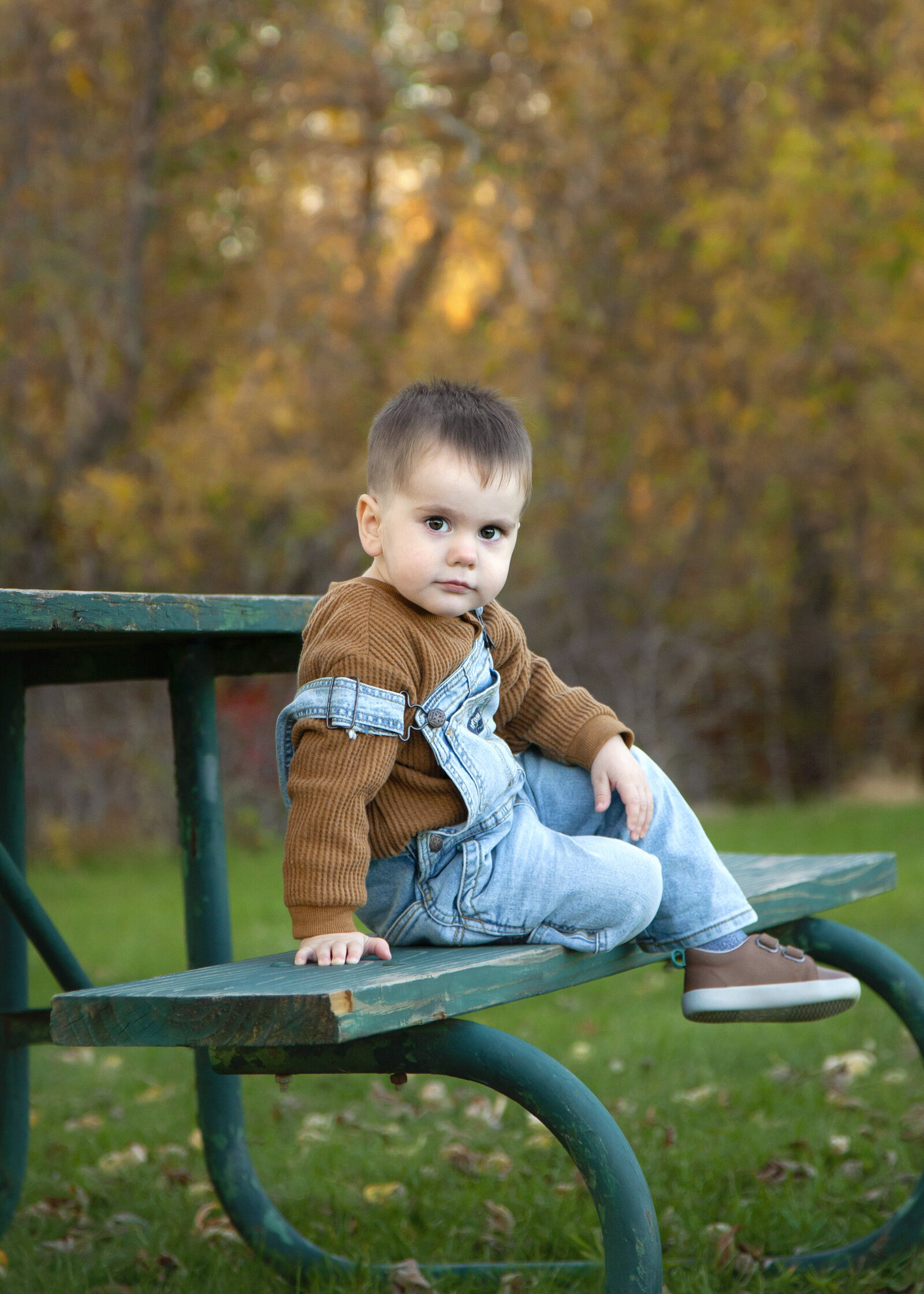 Toddler boy in overalls sitting on picnic bench.