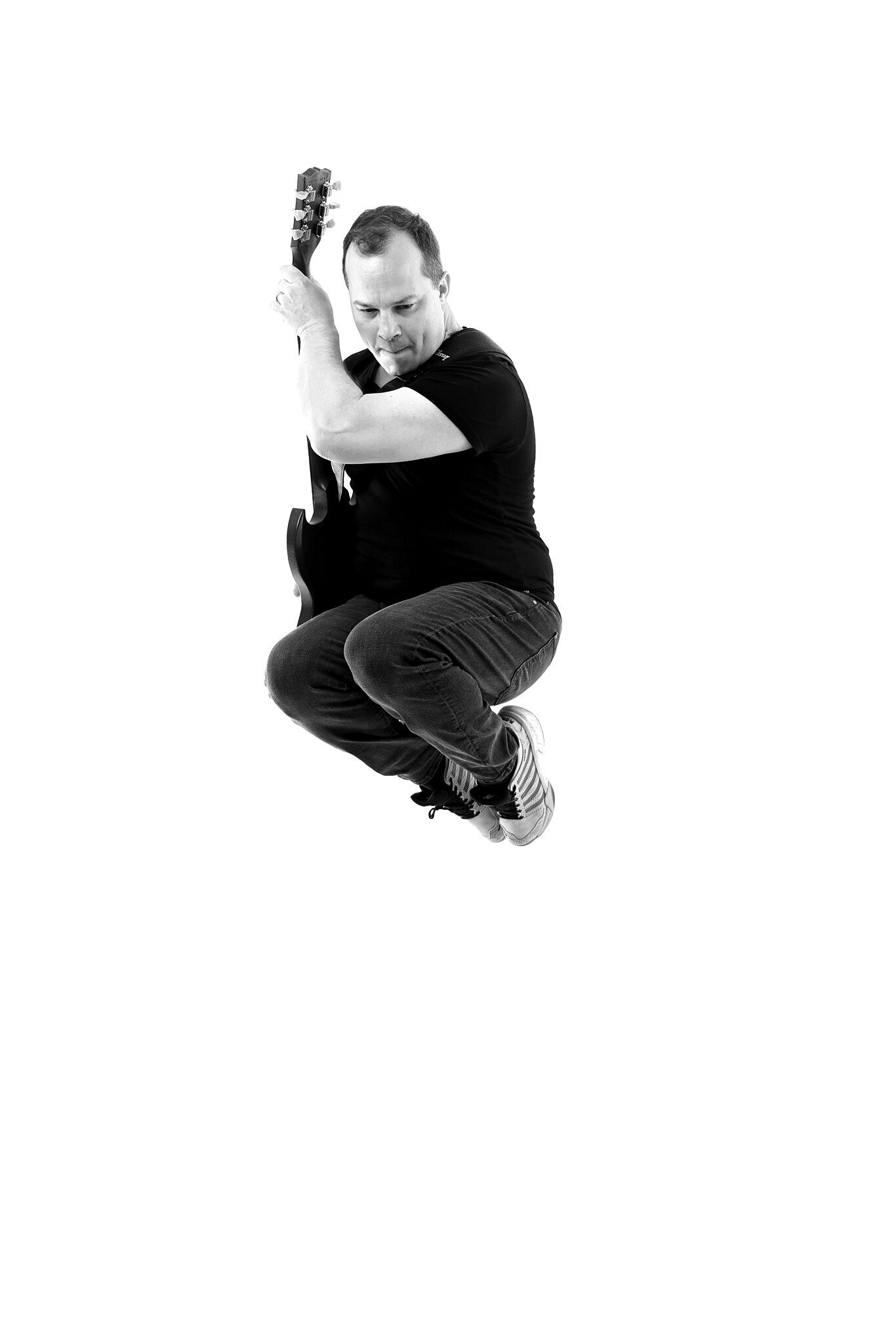 A man wearing a black t-shirt and jeans is jumping in the air while holding an electric guitar. Captured by a Jacksonville band photographer, the high-contrast white background enhances this dynamic music photography shot.