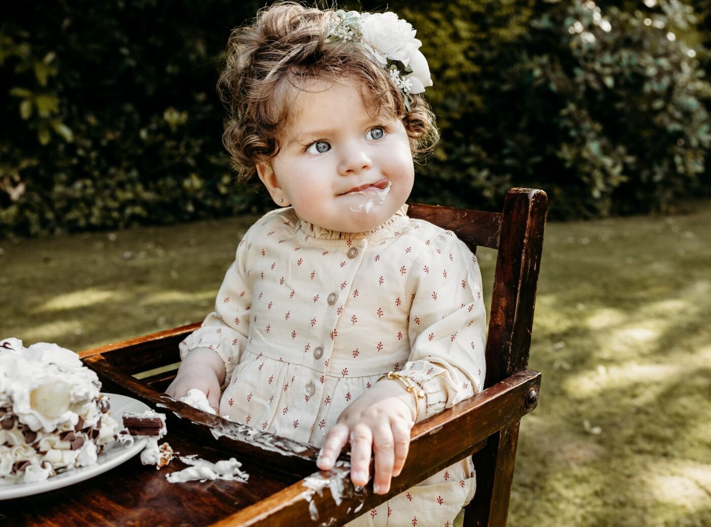 A cute toddler with curly hair and a flower crown sits in a high chair with a messy cake, looking off to the side in a garden by Ruaa
