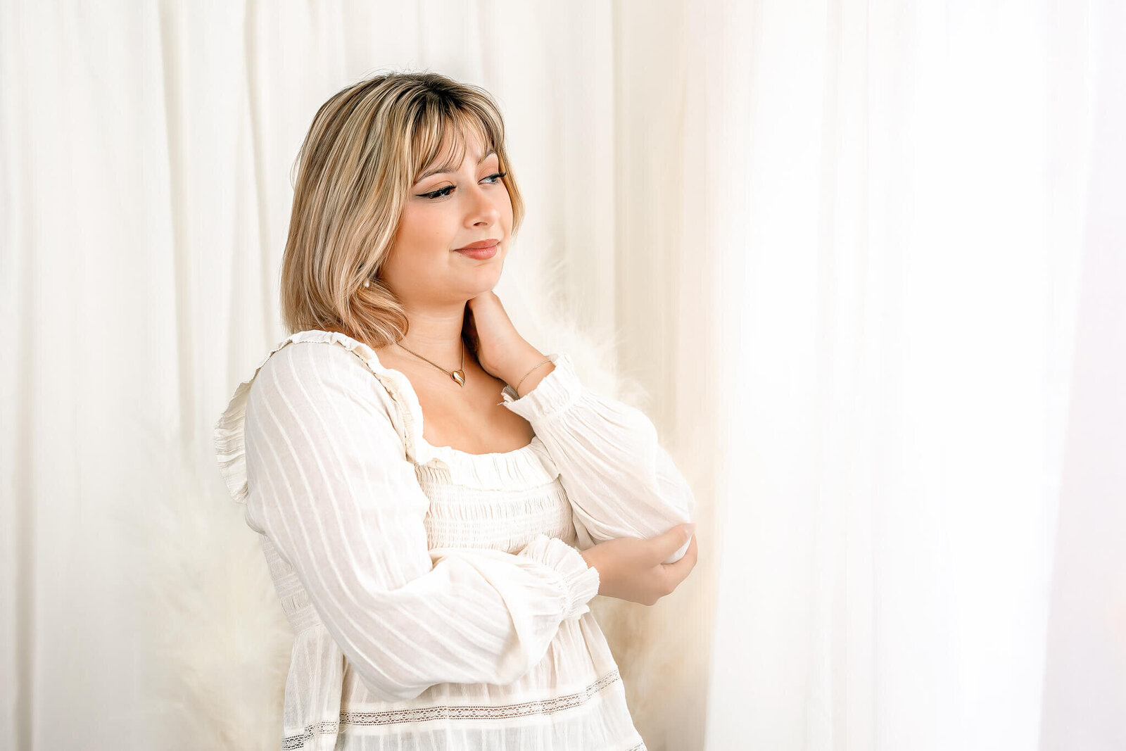 A person with light hair stands against a backdrop of soft, white curtains, wearing a white, textured blouse. They gaze slightly to the side with a relaxed expression, one hand touching their neck.