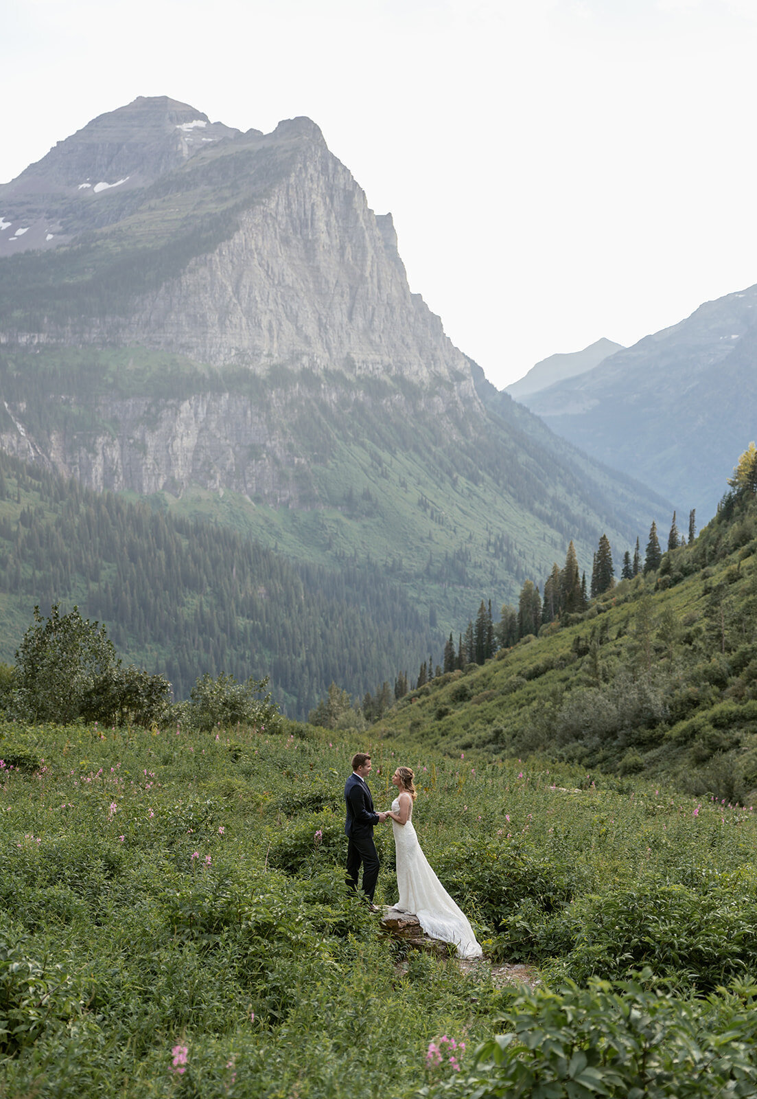A bride and groom stand together in a wildflower meadow surrounded by towering cliffs and pine-covered slopes in Glacier National Park, captured by Sydney Breann Photography during their intimate Montana elopement.