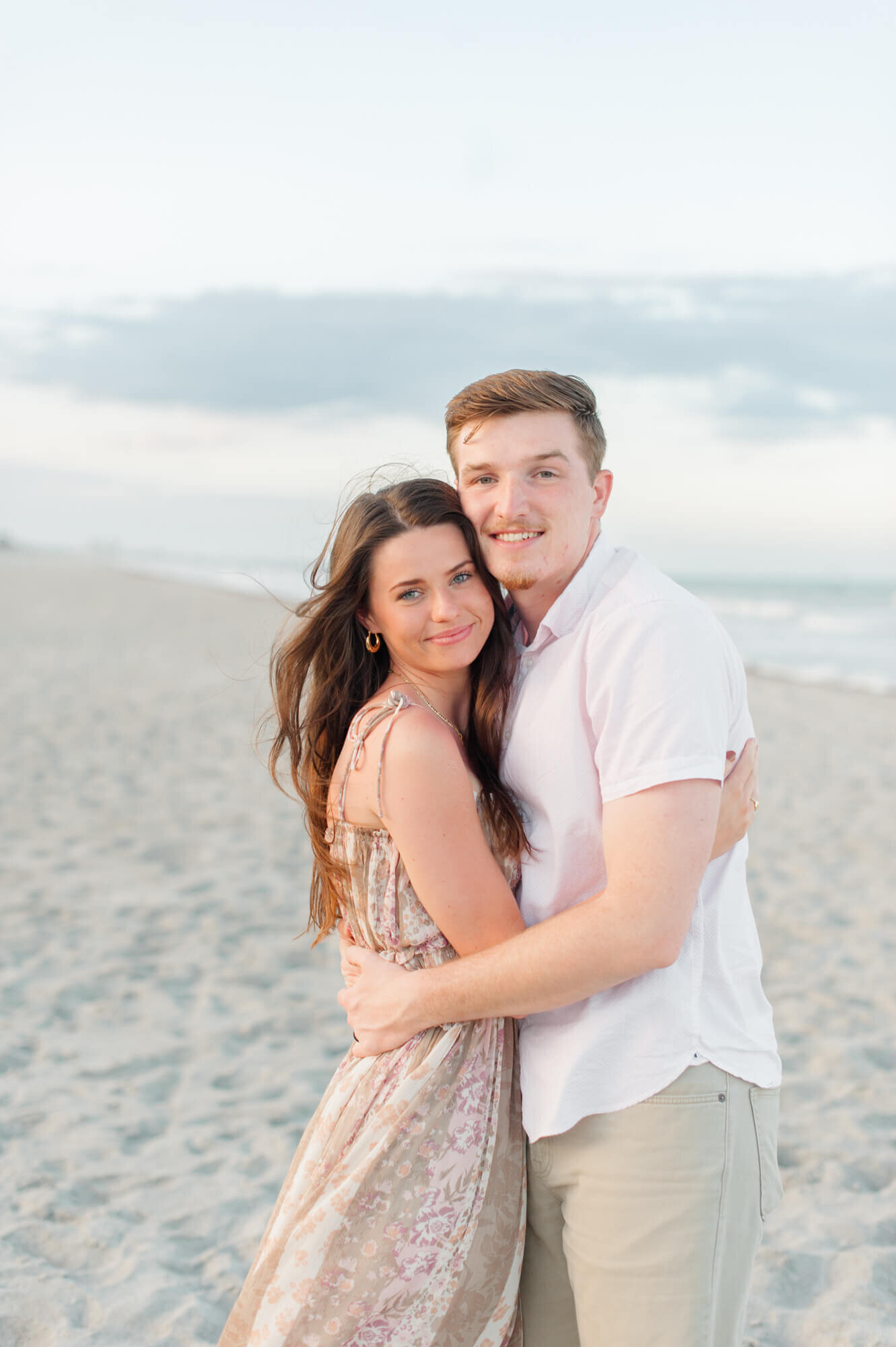 Couple sitting in the dunes with their dog smiling at camera during their engagement session