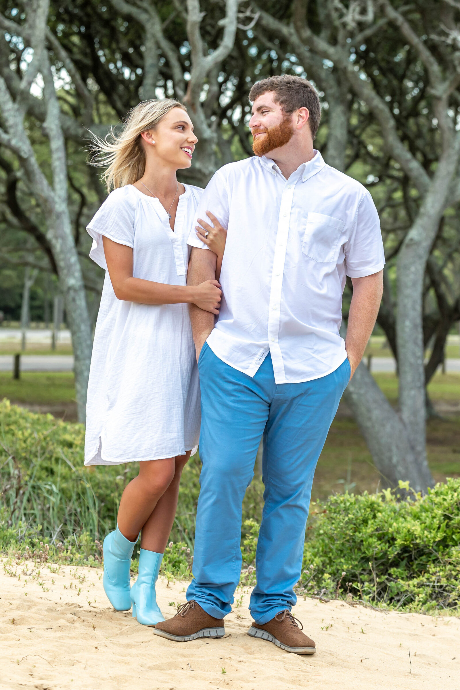 A couple poses affectionately on a sandy path surrounded by a grove of trees. The woman, in a white dress and stylish turquoise boots, gazes at the man, who wears a crisp white shirt and blue pants. This warm, natural setting embodies the timeless charm of North Carolina engagement photography. 