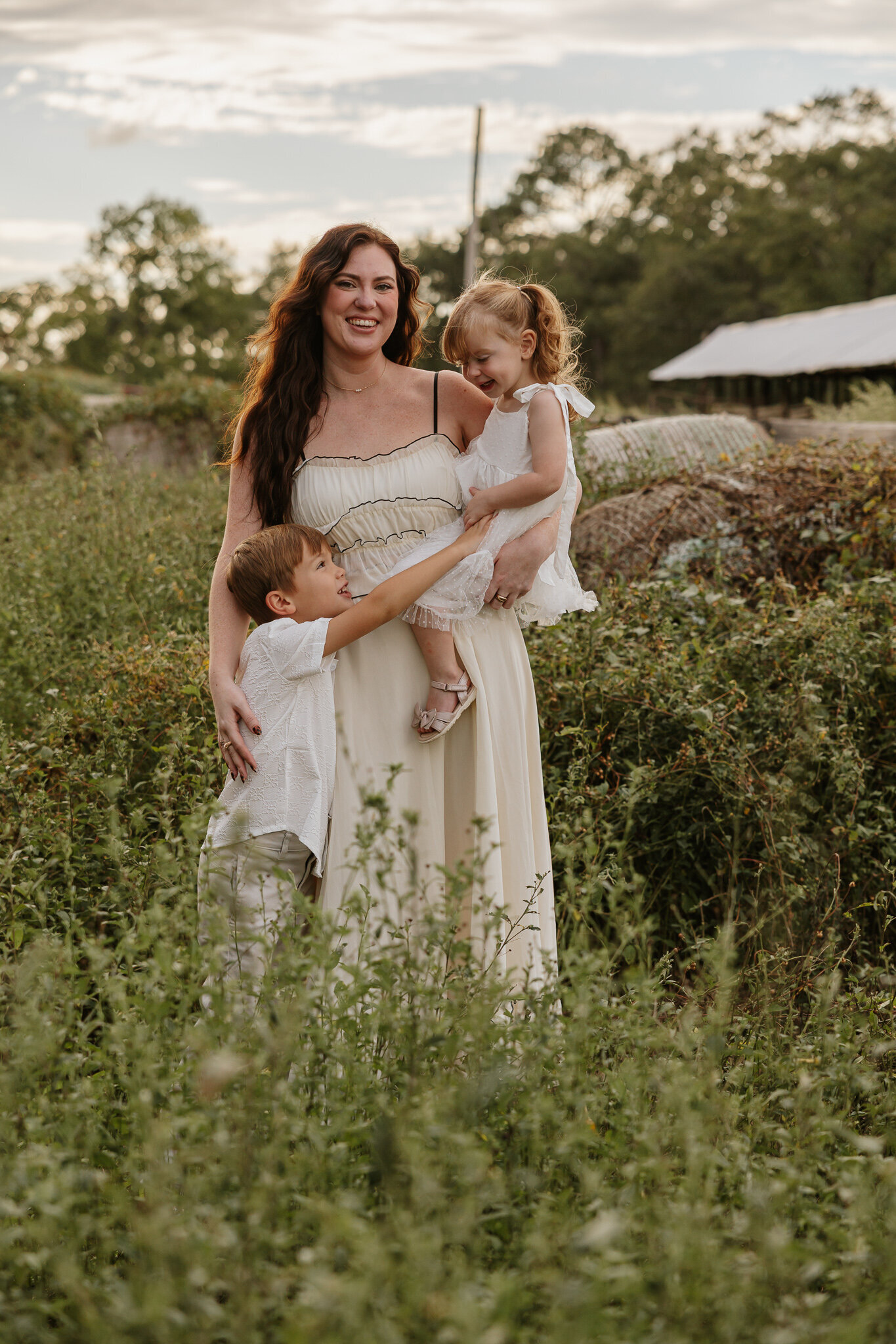 Mother holding her daughter while the older brother tickles his sister during a golden hour family session in a fam field in Aiken SC.