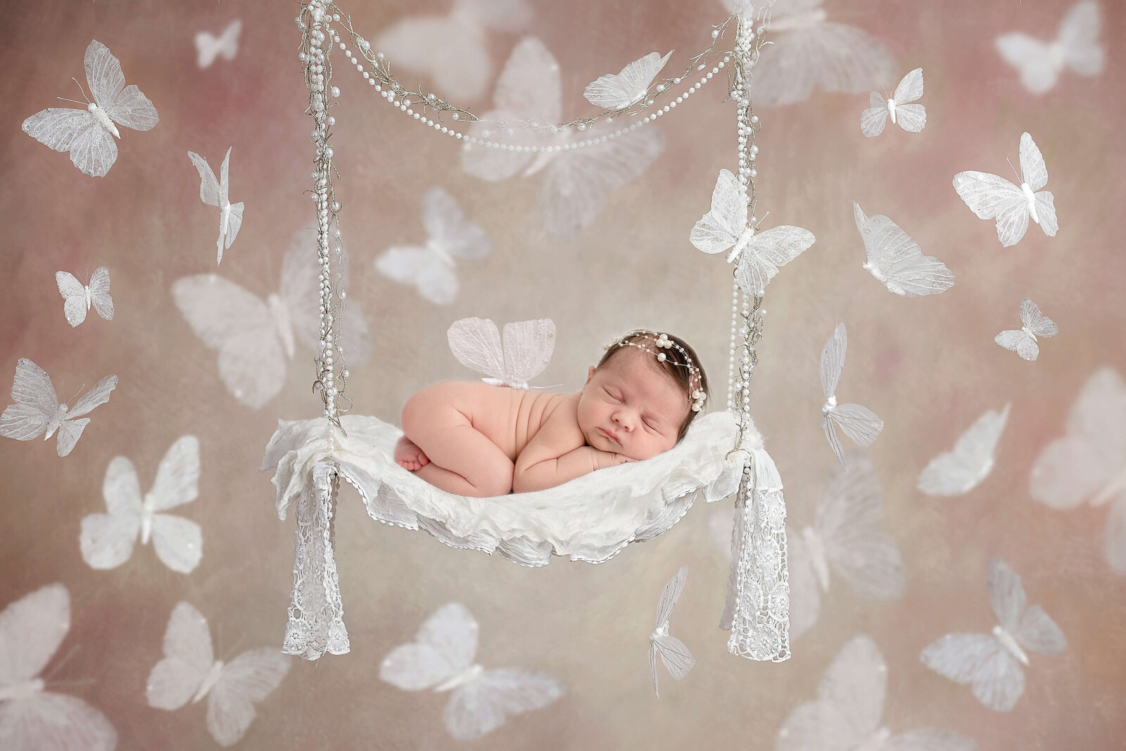 Newborn baby sleeping on a swing surrounded by white butterflies wearing a pearl headband