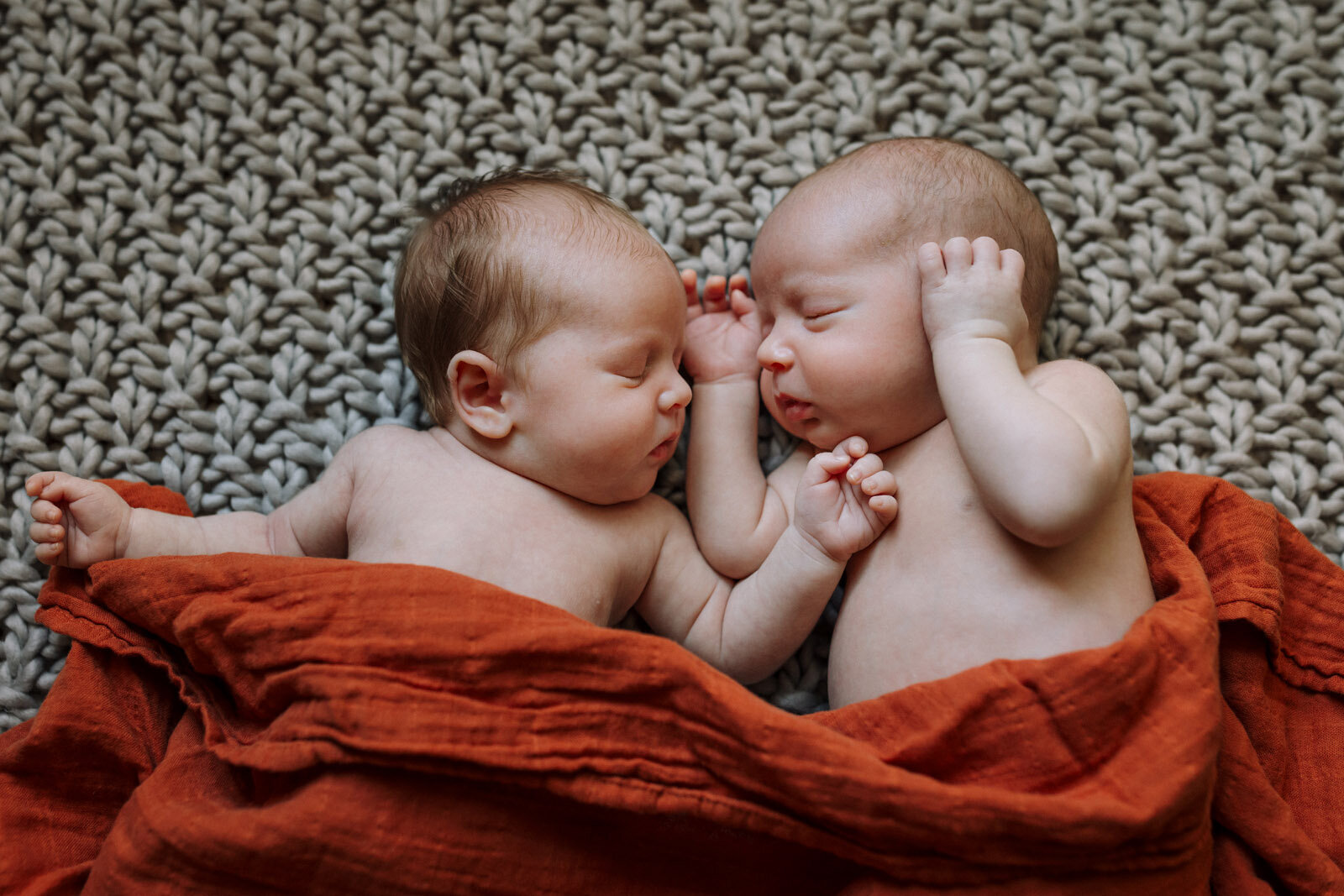 newborn twins sleeping facing each other on textured blanket with orange swaddle blanket draped over them
