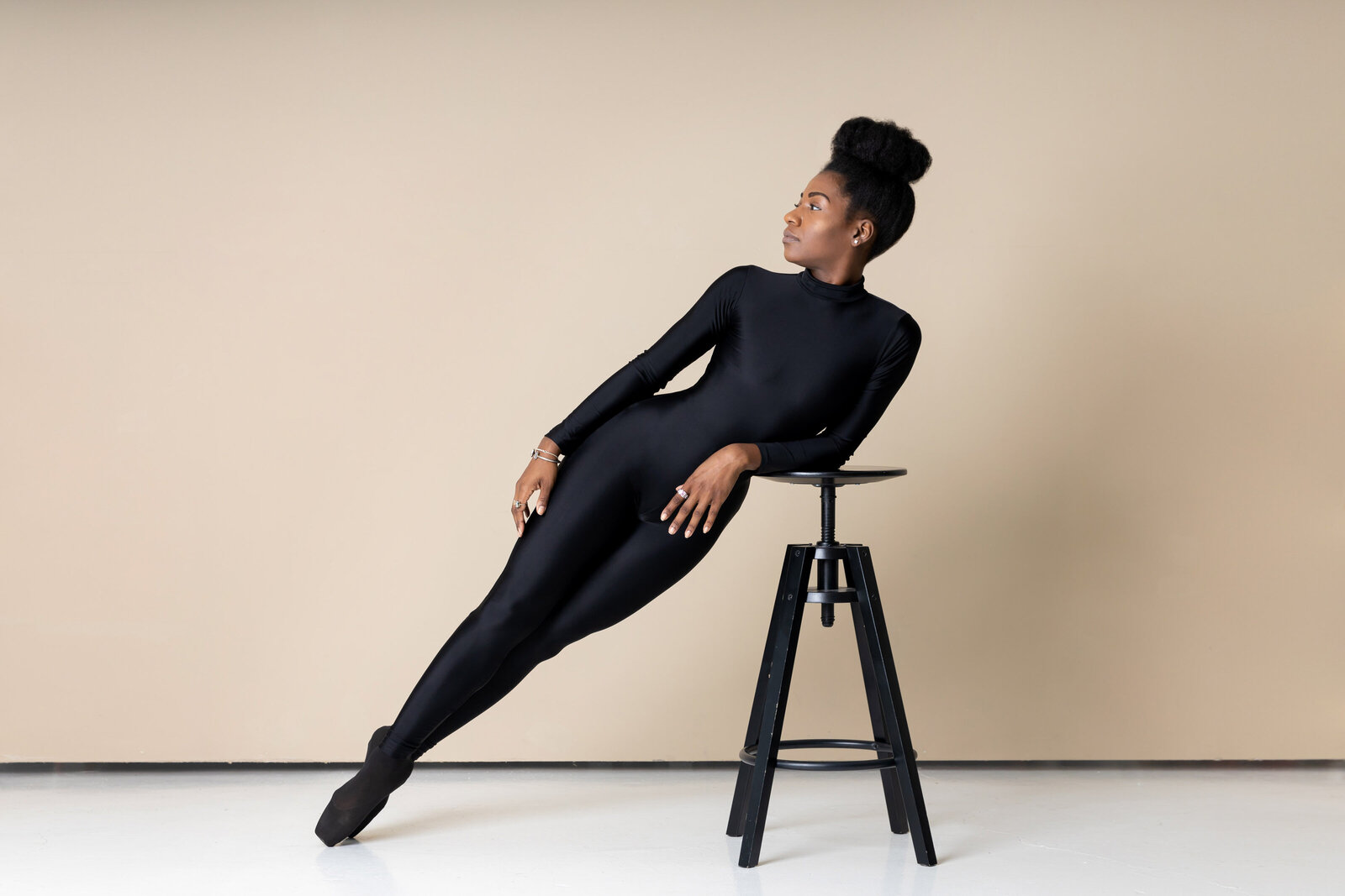 black ballerina in black unitard and pointe shoes leaning on a black stool