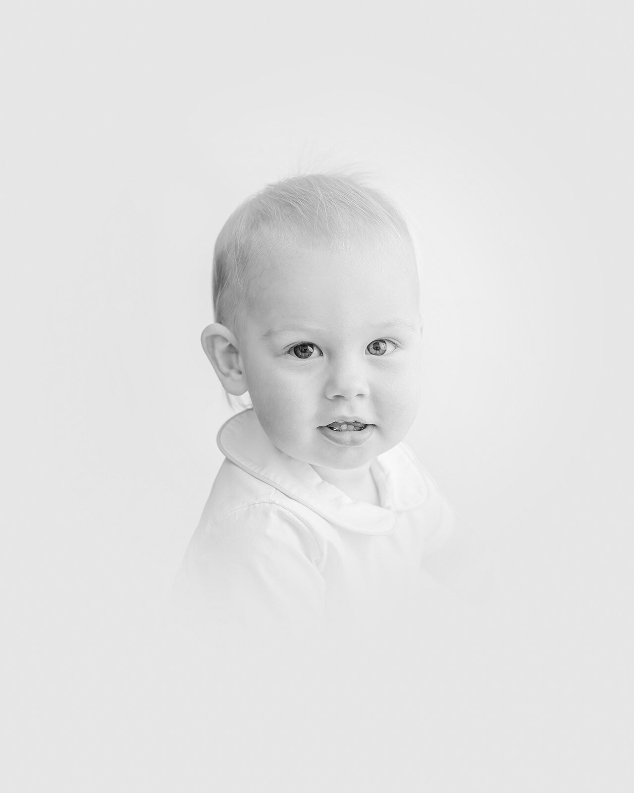 A timeless portrait captures a baby with blue eyes and light hair, clad in a white shirt, gazing into the camera against a simple, light backdrop.