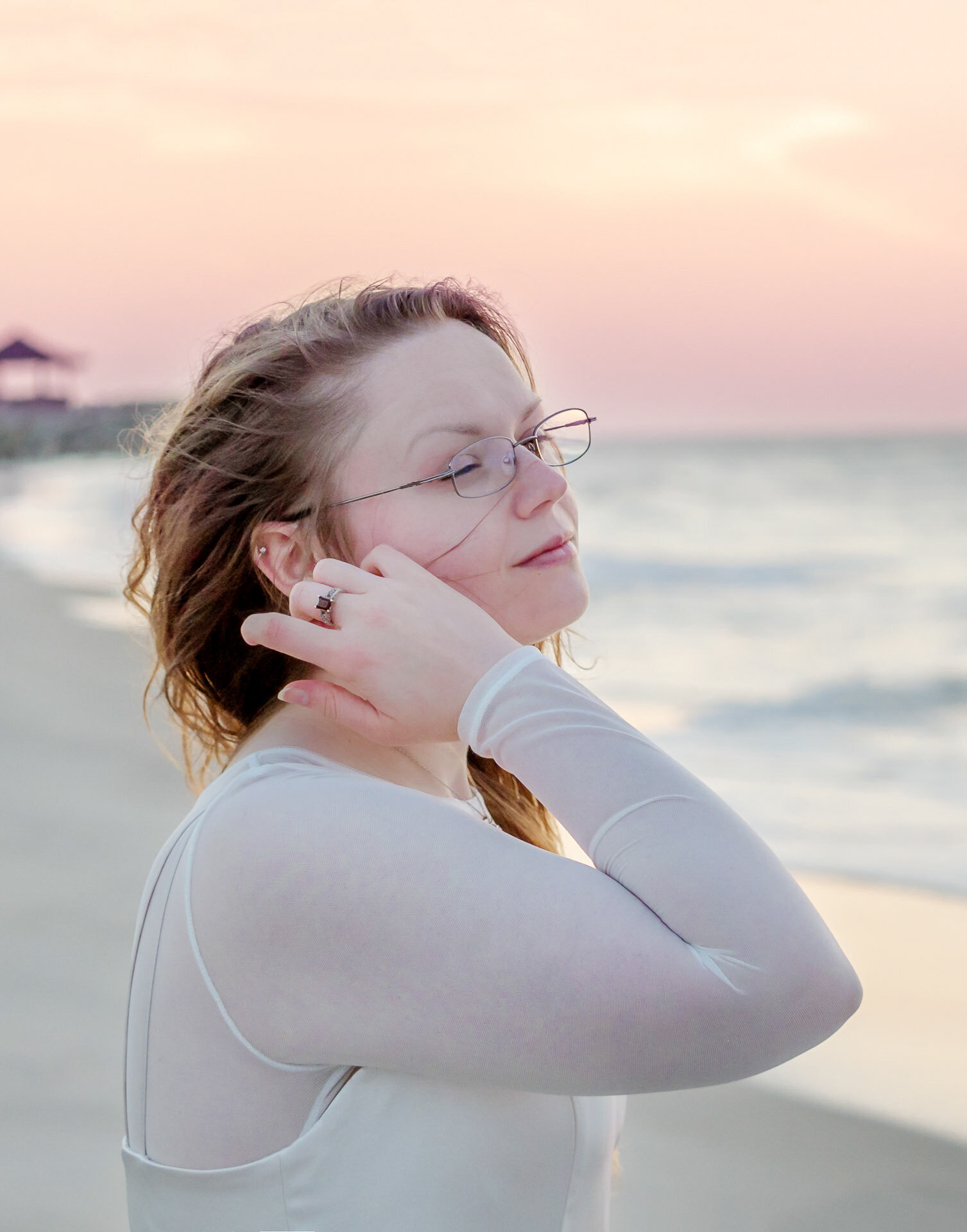 A college senior with glasses gently touching her hair, standing on the beach at sunset in North Carolina. This serene portrait captures the elegance of senior photography, ideal for those seeking artistic and natural beach photos in Wilmington, NC.