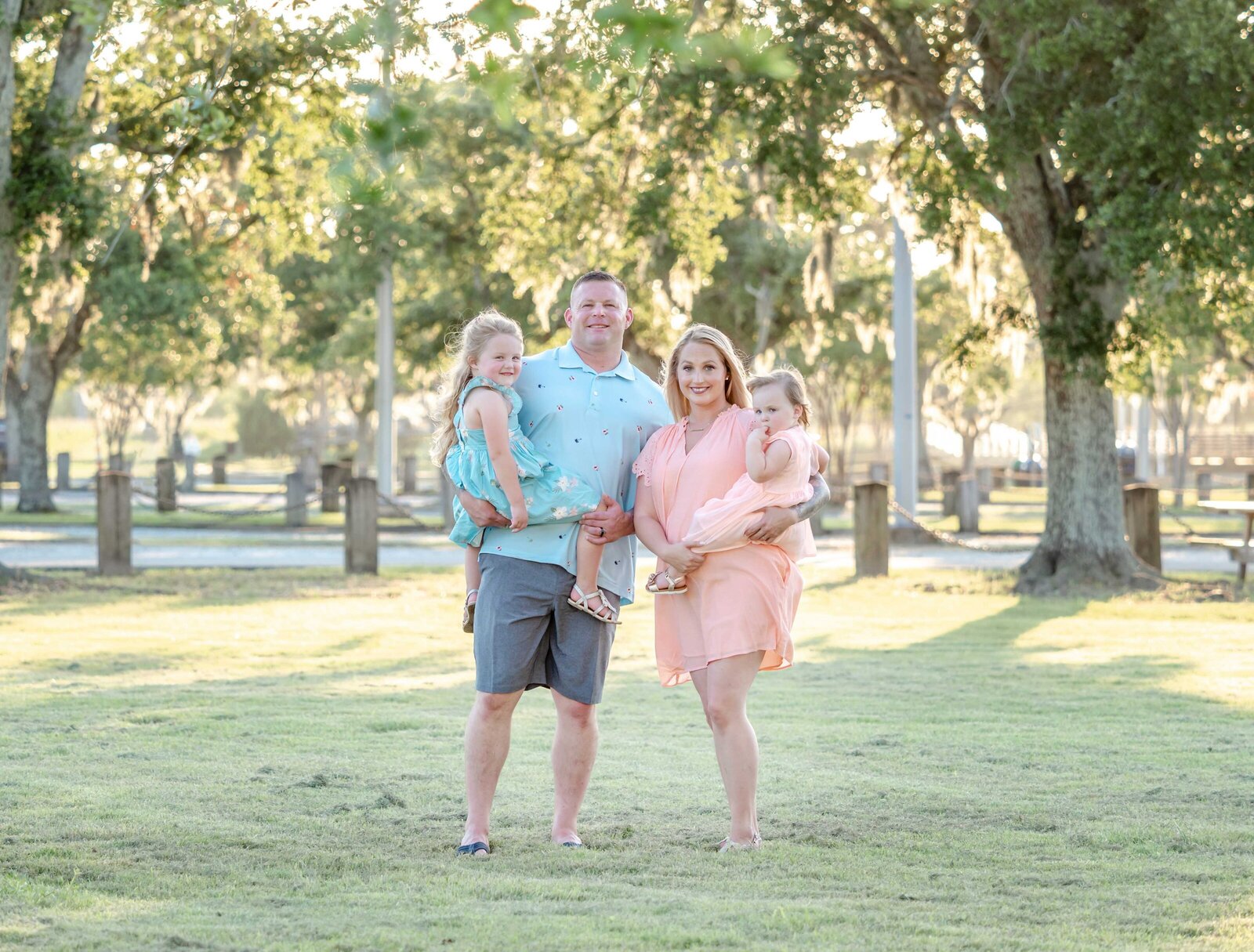 A family of four stands in a sunlit park near Wilmington, NC. The parents hold their young daughters, all dressed in pastel outfits, surrounded by lush greenery. This image highlights affordable family photography in Wilmington, NC, capturing warm family bonds.