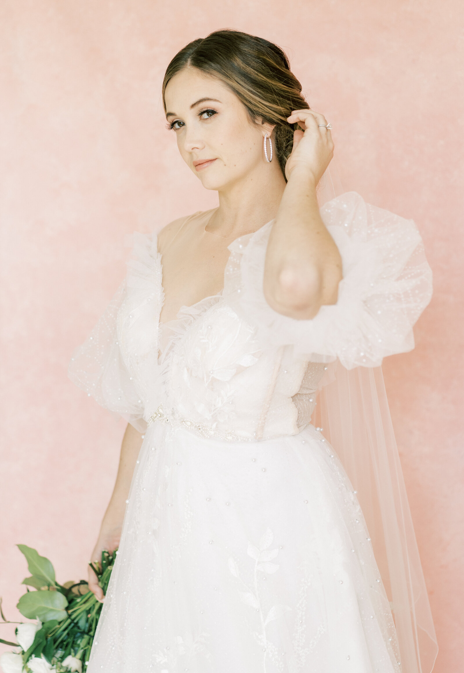 Portrait of a bride in a white beaded wedding gown with pink backdrop and bouquet of flowers.