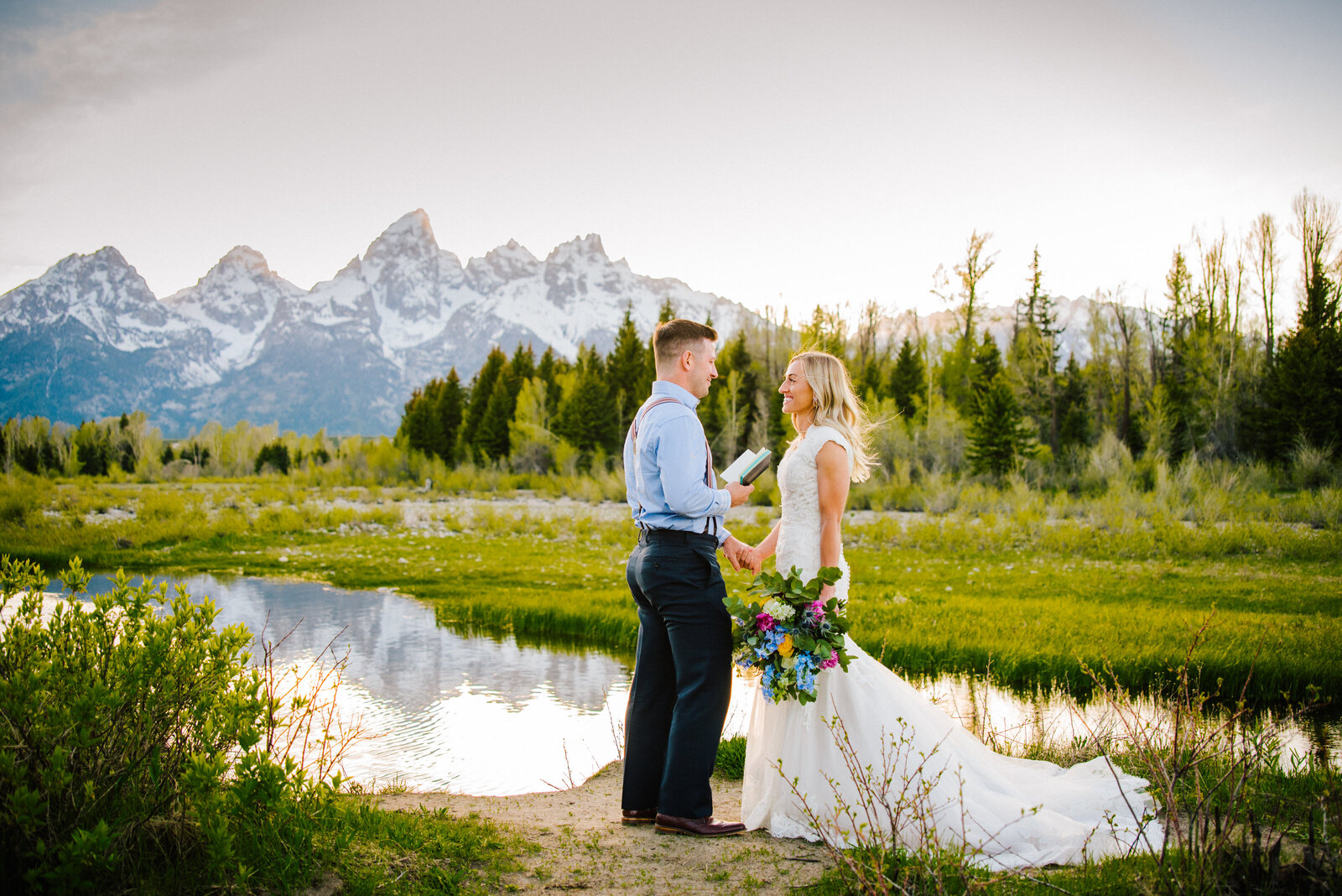 tender and emotional vow reading  at jackson hole elopement by jackson hole wedding phtoographer. Bride and groom hold hands as they read their wedidng vows to each other by the waters edge with the tetons behind him.